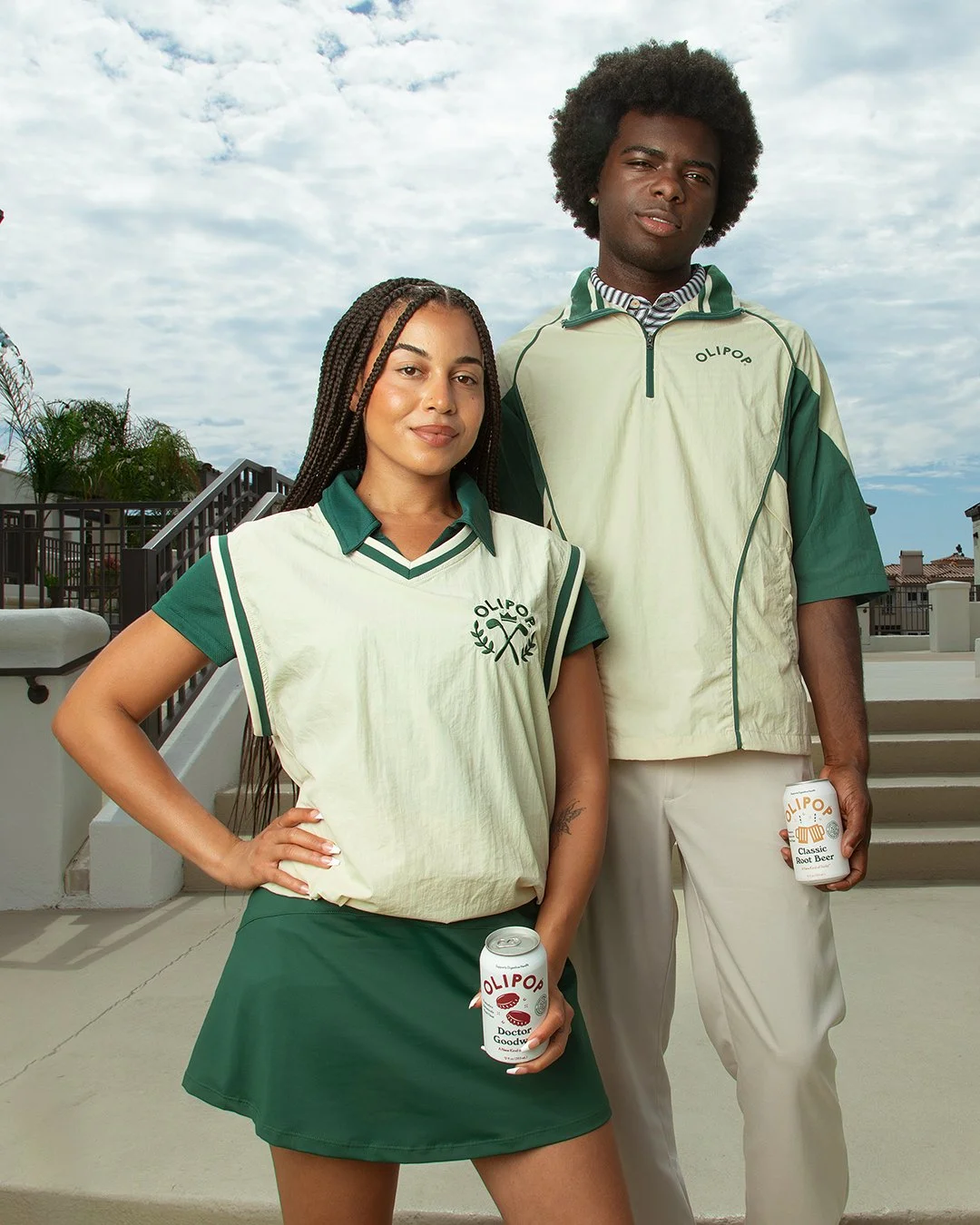 Two young adults standing outdoors on a bright day, holding cans of Olipop drink, with a sky and rooftops in the background.