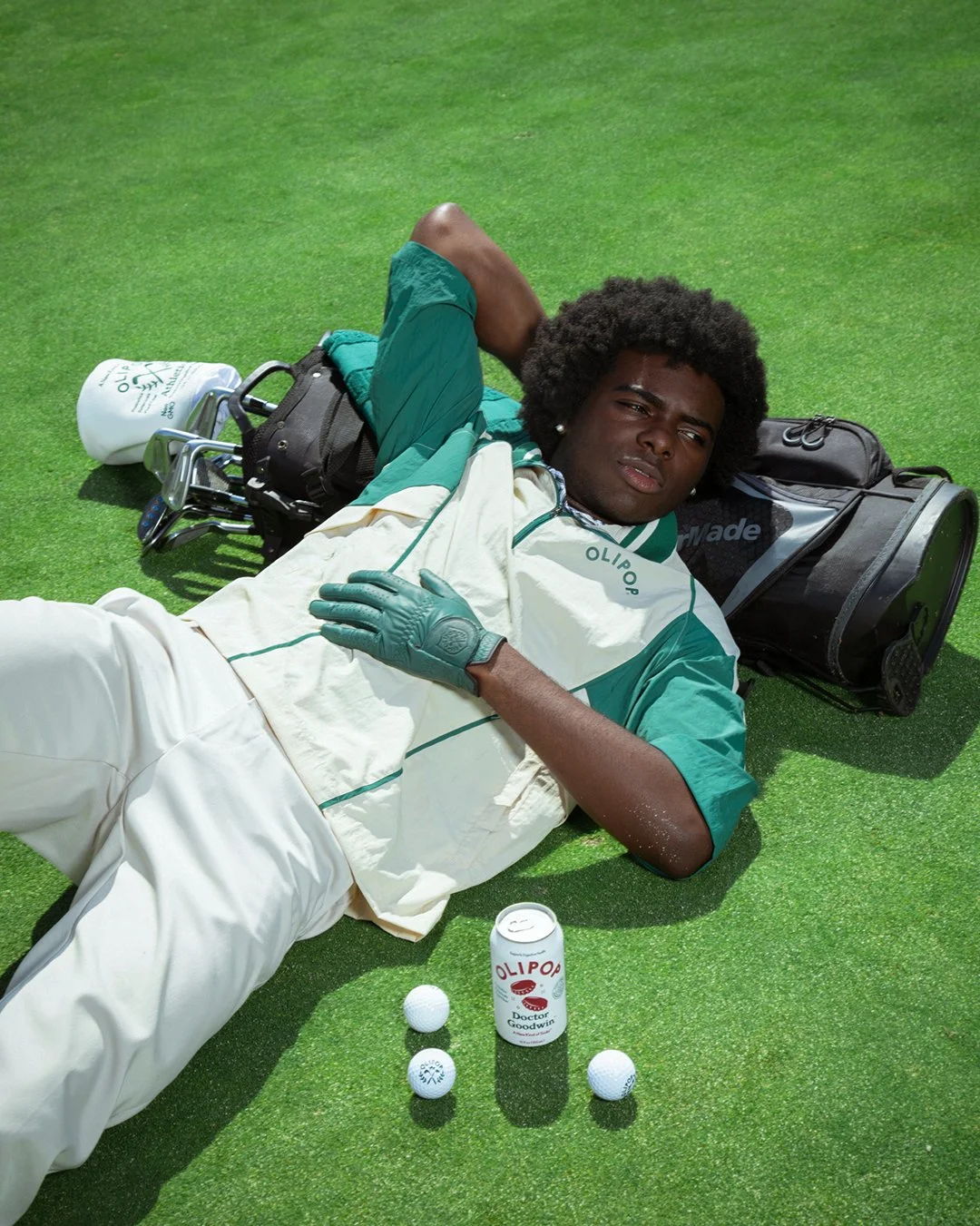 A male golfer lying on artificial grass, wearing golf gloves, a white and green golf shirt, and cream-colored pants. There are three golf balls and a drink can with the label 'OliPop' and 'Doctor Goodwin', nearby.