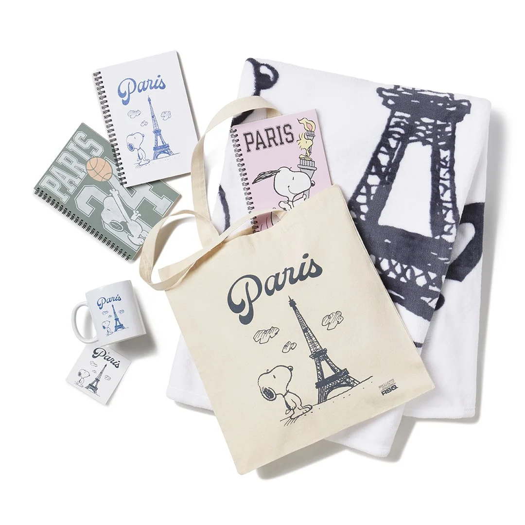 Collection of Paris-themed Snoopy souvenirs including notebooks, mugs, a tote bag, and a towel with Eiffel Tower and Snoopy illustrations.