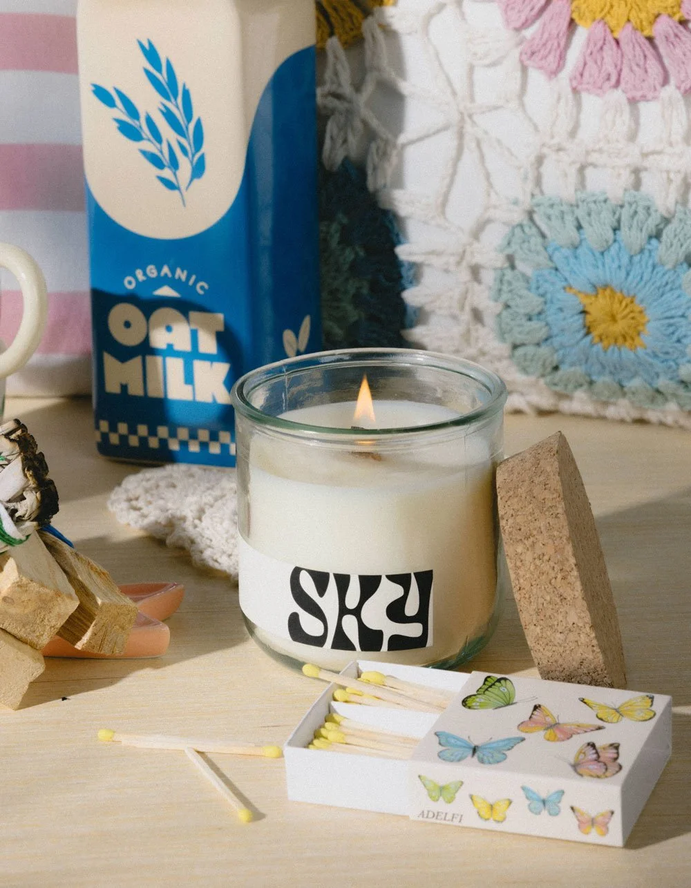 A lit candle in a glass container with a black and white label, a box of butterfly-themed matches, a corkboard, a carton of organic oat milk, and a crocheted pillow with pastel flowers and leaves on a wooden surface.