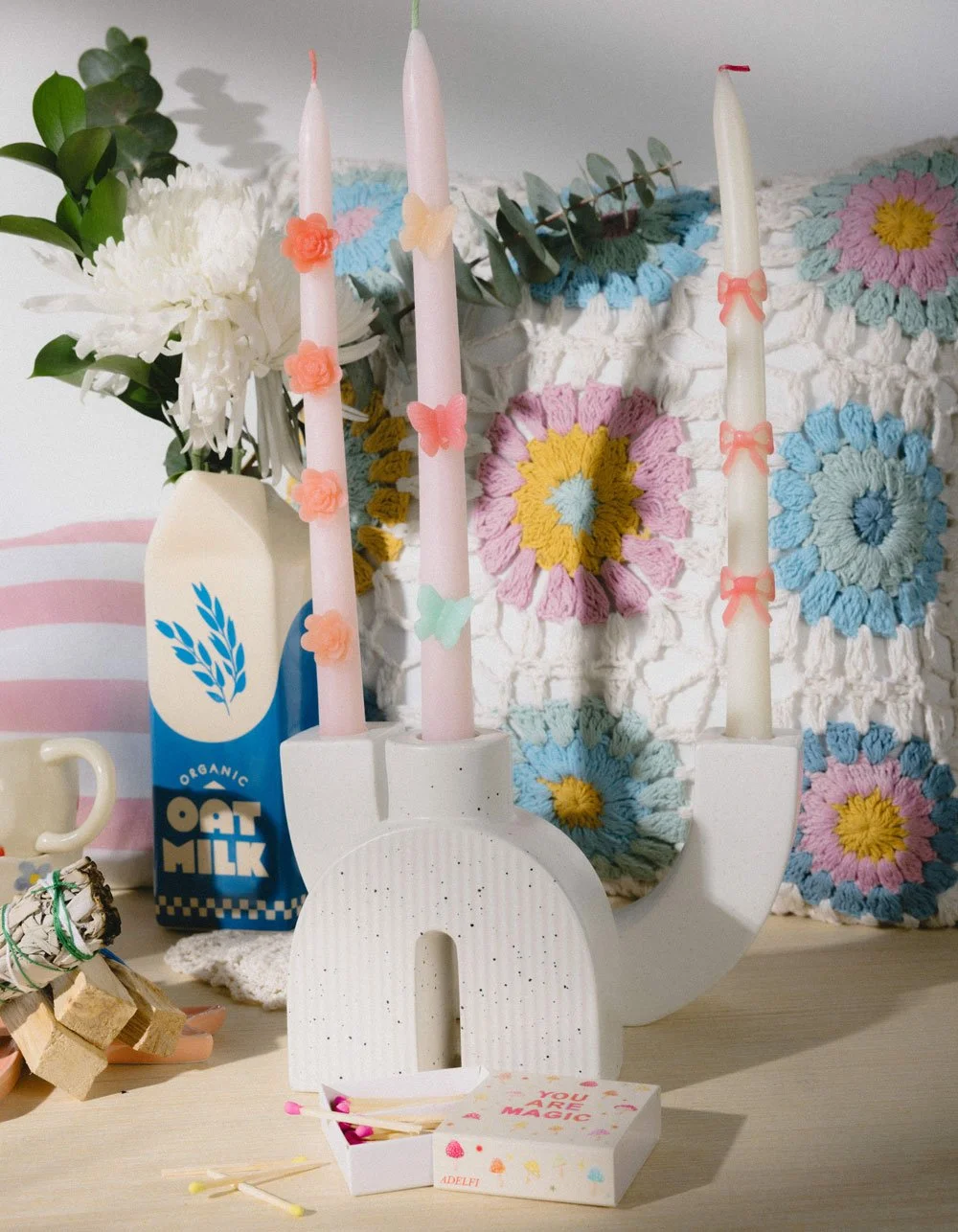 Decorative scene including a white letter 'U' candle holder with three pink candles decorated with small bows and flowers, a carton of organic oat milk, a small white cup, a bundle of sage sticks, a box of matches labeled 'You Are Magic', a white vas