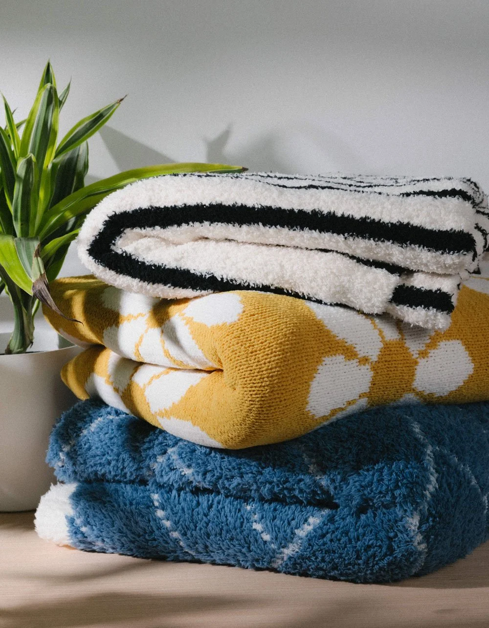 A stack of three folded towels with a plant in a white pot to the left. The towels are of different colors and patterns: a black and white striped towel on top, a yellow and white patterned towel in the middle, and a solid blue towel at the bottom.