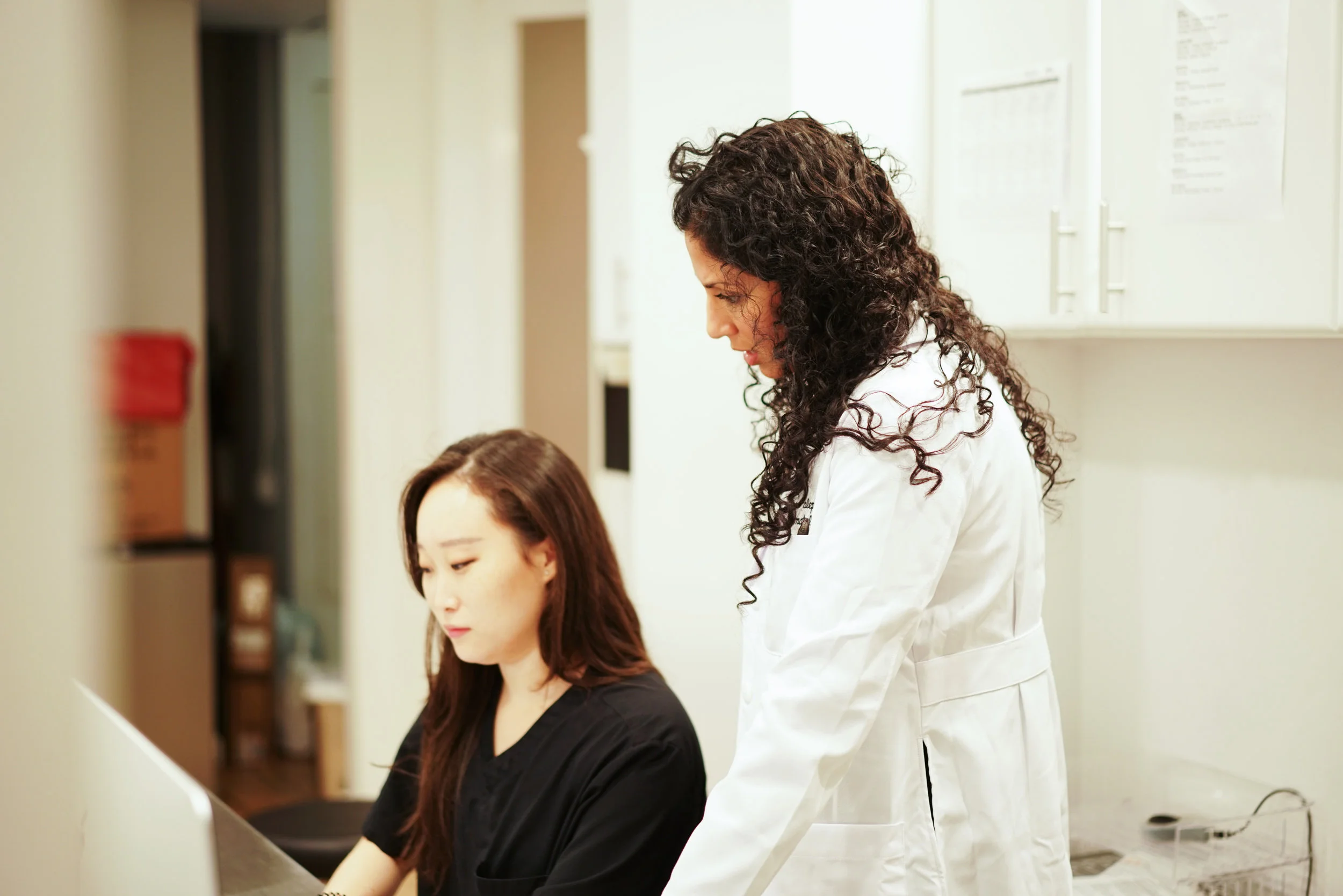 Image of a doctor in an office standing up next to an assistant who’s sitting down next to her