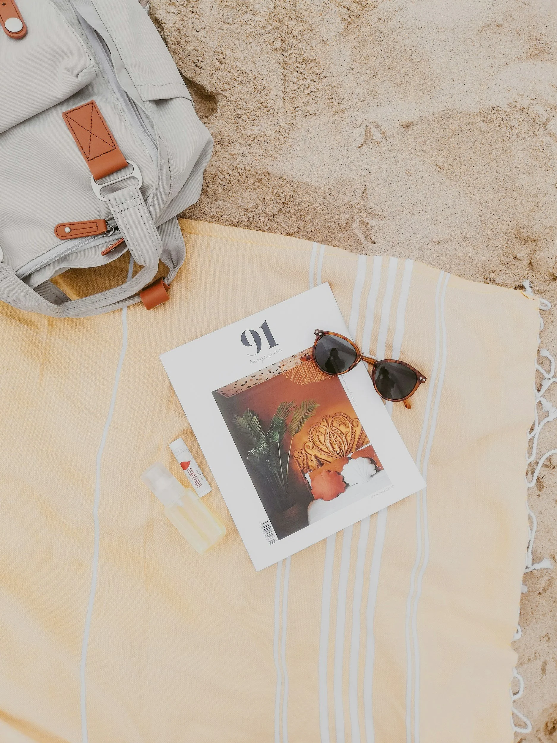 Beach scene with a gray backpack, a yellow and white striped towel, a magazine, sunglasses, a small bottle of sunblock, and lip balm on the sand.
