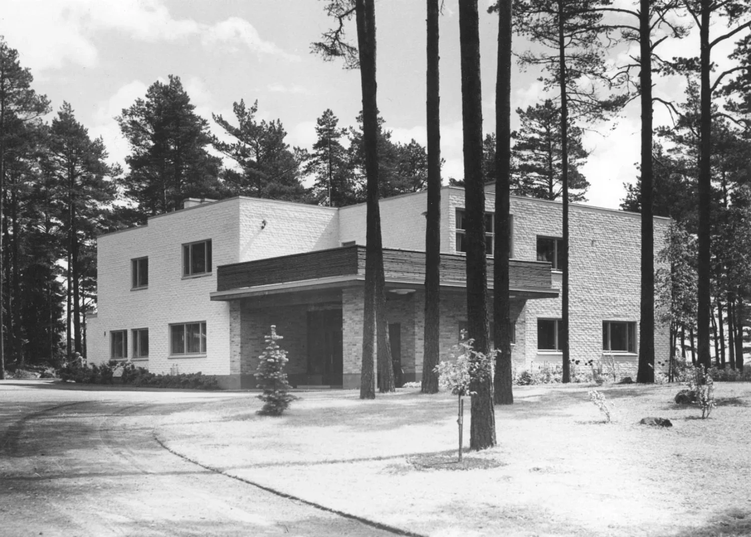 Mustavalkokuva Kantolasta 1930-luvun lopulta. Sisäänkäynnille vie kaareutuva hiekkatie ja vaaleaa rakennusta ympäröi erilaiset matalat itutukset. Männyt etualalla peittävät hieman näkymää rakennukselle.