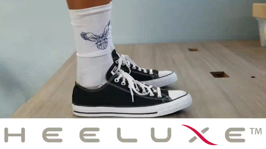 A person wearing black low-top Converse-style trainers and white socks with an owl graphic stands on a wooden platform. The Heeluxe™ logo appears beneath the image