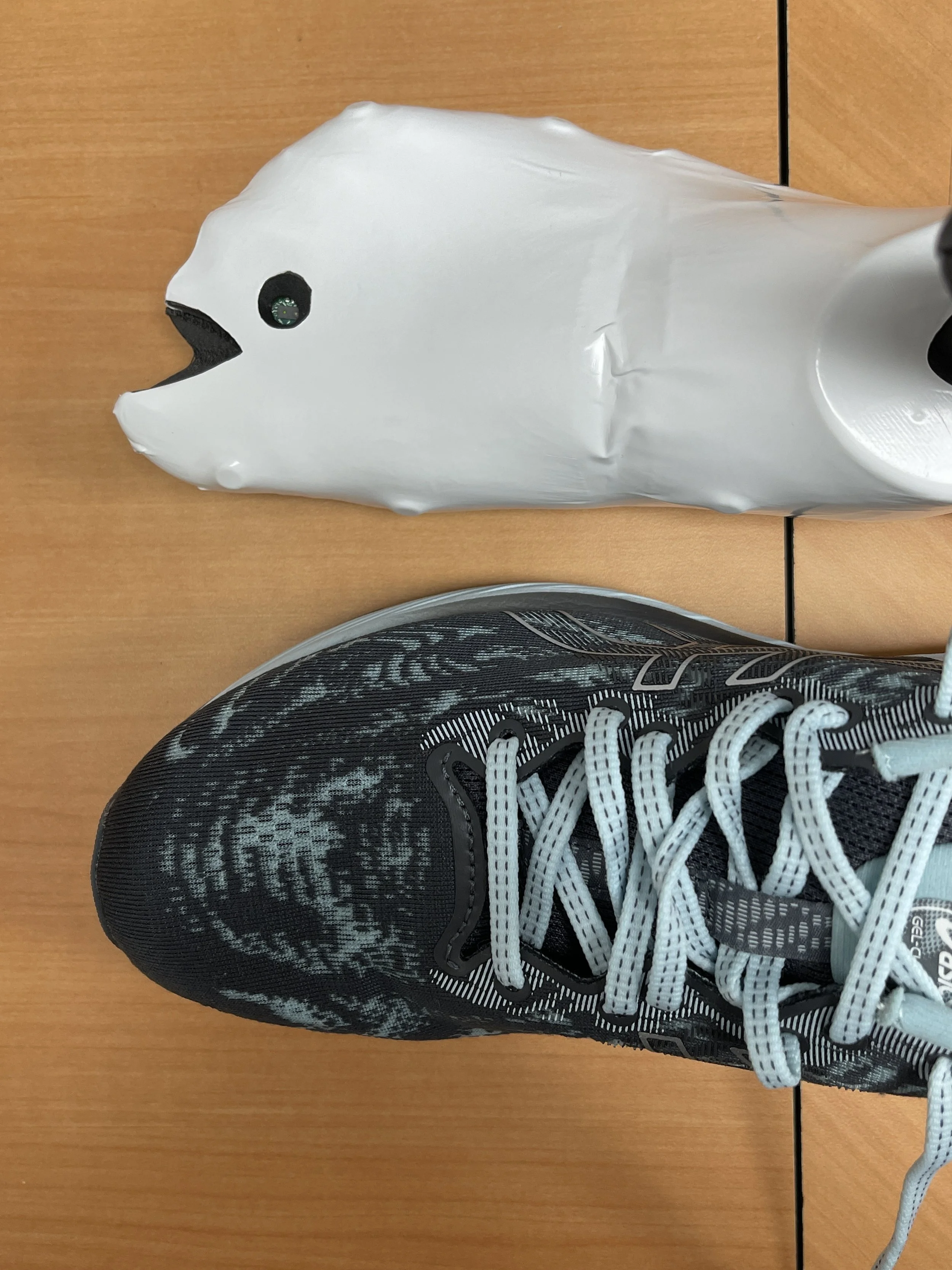 A top-down view of a running shoe next to a white, foot-shaped SmartLast testing device. The device has a small circular sensor near the toe area and resembles a wrapped mould of a foot. Both objects rest on a wooden surface.