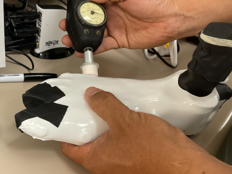 A technician holds a white SmartLast foot-shaped device while using a handheld gauge to calibrate one of its embedded sensors. Black tape reinforces part of the device, and cables and equipment are visible in the background.