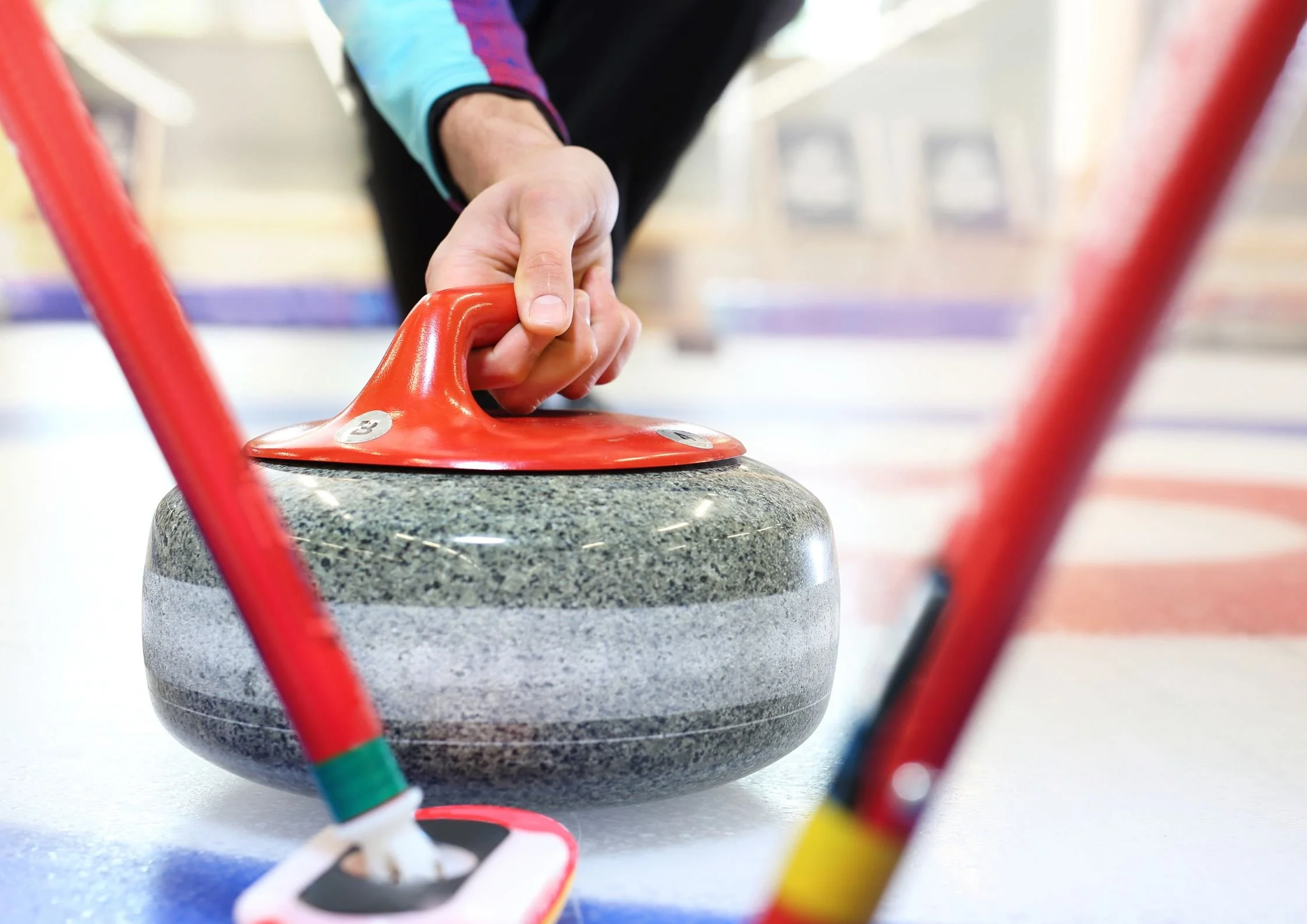 Curler gripping the handle of a granite curling stone on the ice, with sweepers and brushes visible in the foreground during competitive play.