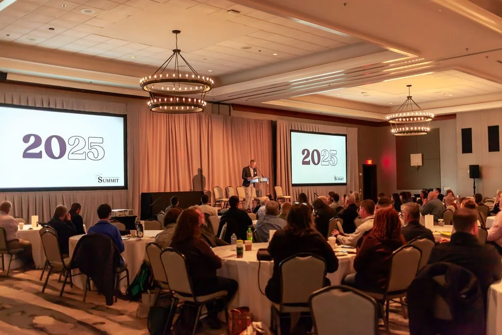 A large group of people are in a dimly lit conference room while a man speaks on stage