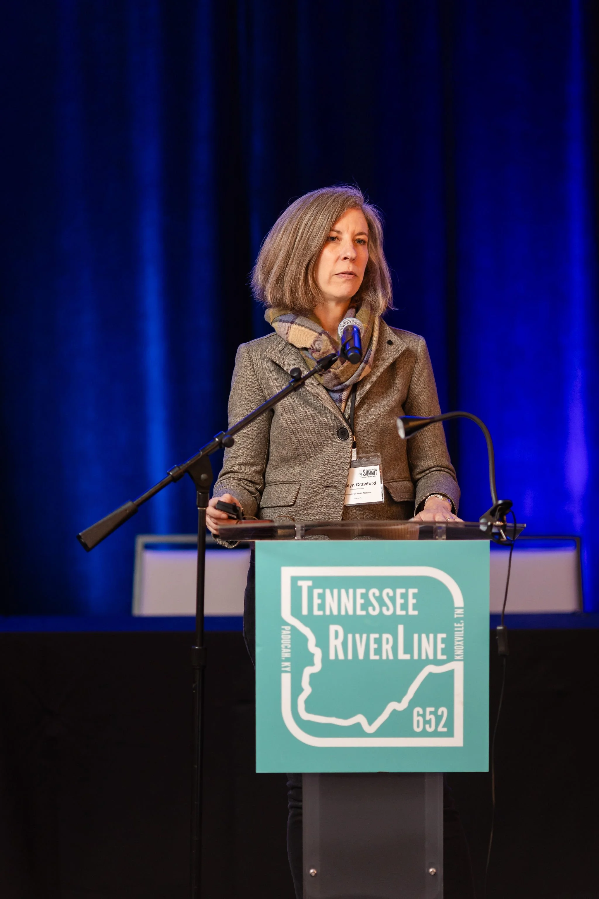 A woman wearing a brown blazer and scarf stand behind a lectern displaying the Tennessee RiverLine logo.