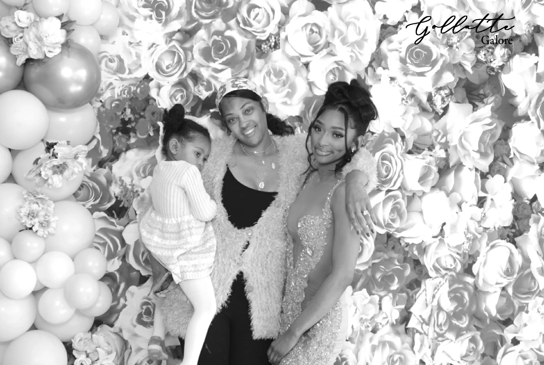 A woman in a sparkly dress, a woman in a fuzzy cardigan, a young girl, and another woman standing in front of a floral backdrop with balloons. The woman in the dress is smiling, and the young girl is looking at her.