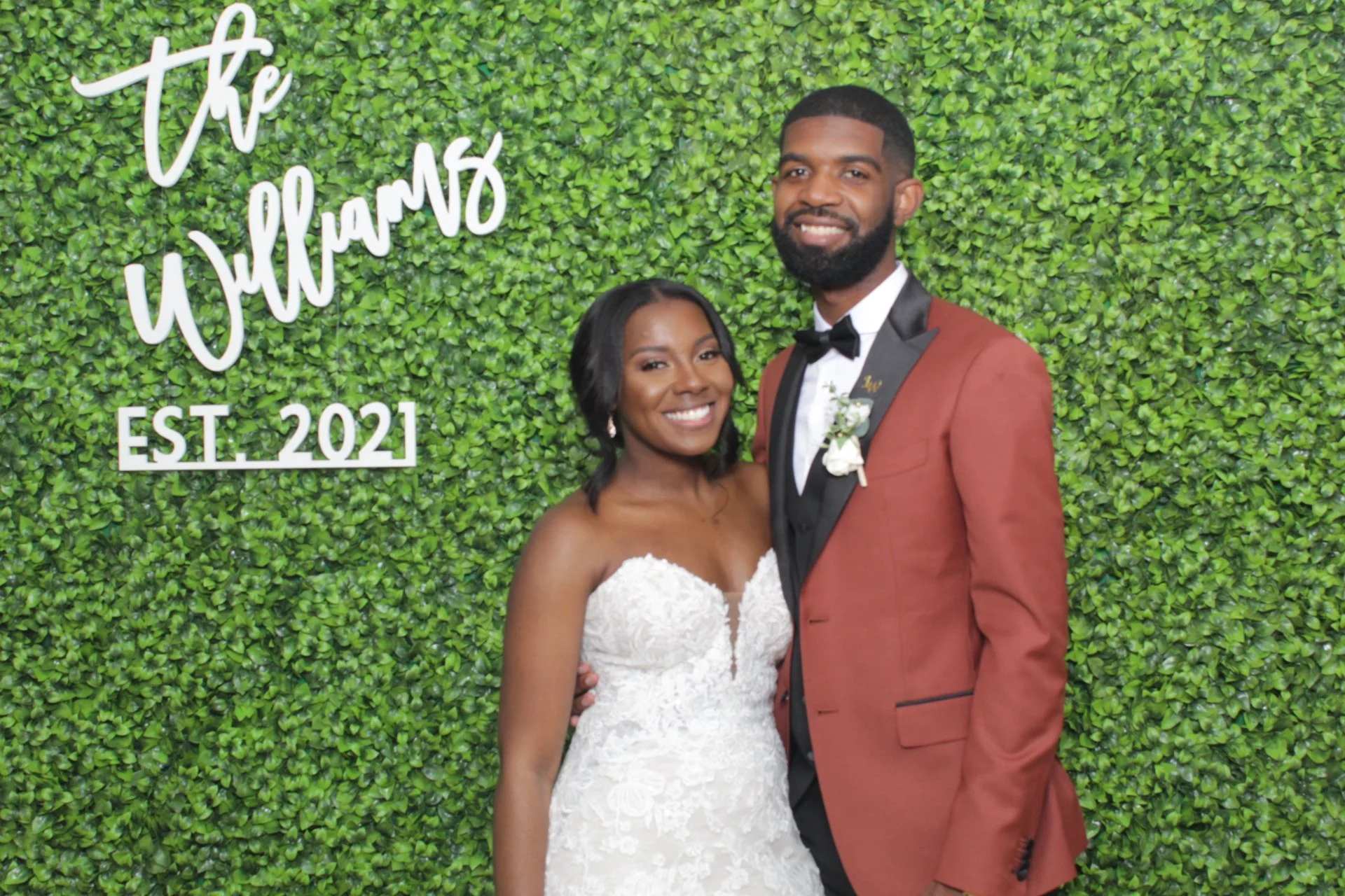 A happy couple in wedding attire standing in front of a green leafy wall with white cursive text that reads 'The Gillams' and 'Est. 2021'.