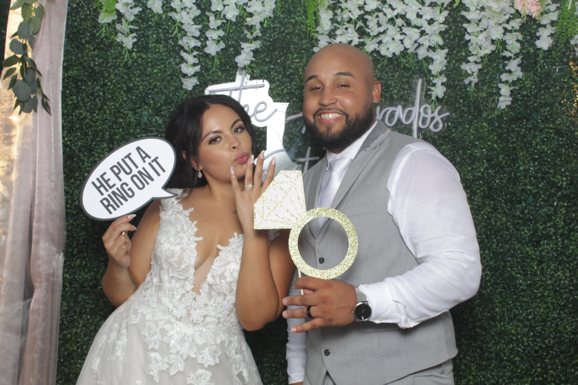 A bride and groom posing together at their wedding. The bride is holding a speech bubble sign that says 'HE PUT A RING ON IT' and is making a kissy face. The groom is smiling and holding a large glittery ring prop with the number 5. The background is a green leafy wall with decorative white flowers and neon signs.
