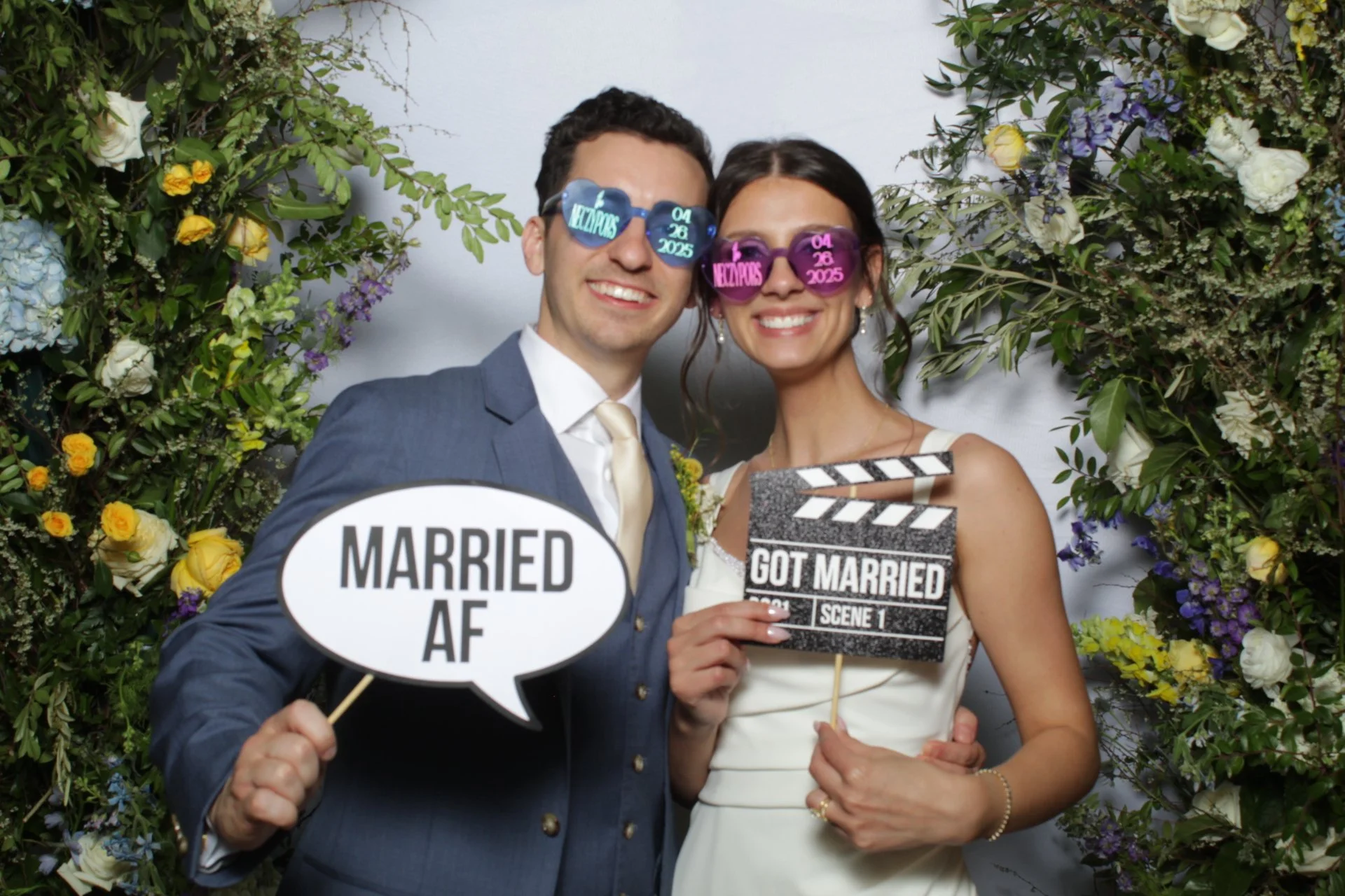 A smiling man and woman at their wedding surrounded by flowers, holding signs that say 'Married A.F.' and 'Got Married,' wearing sunglasses with date and location details.