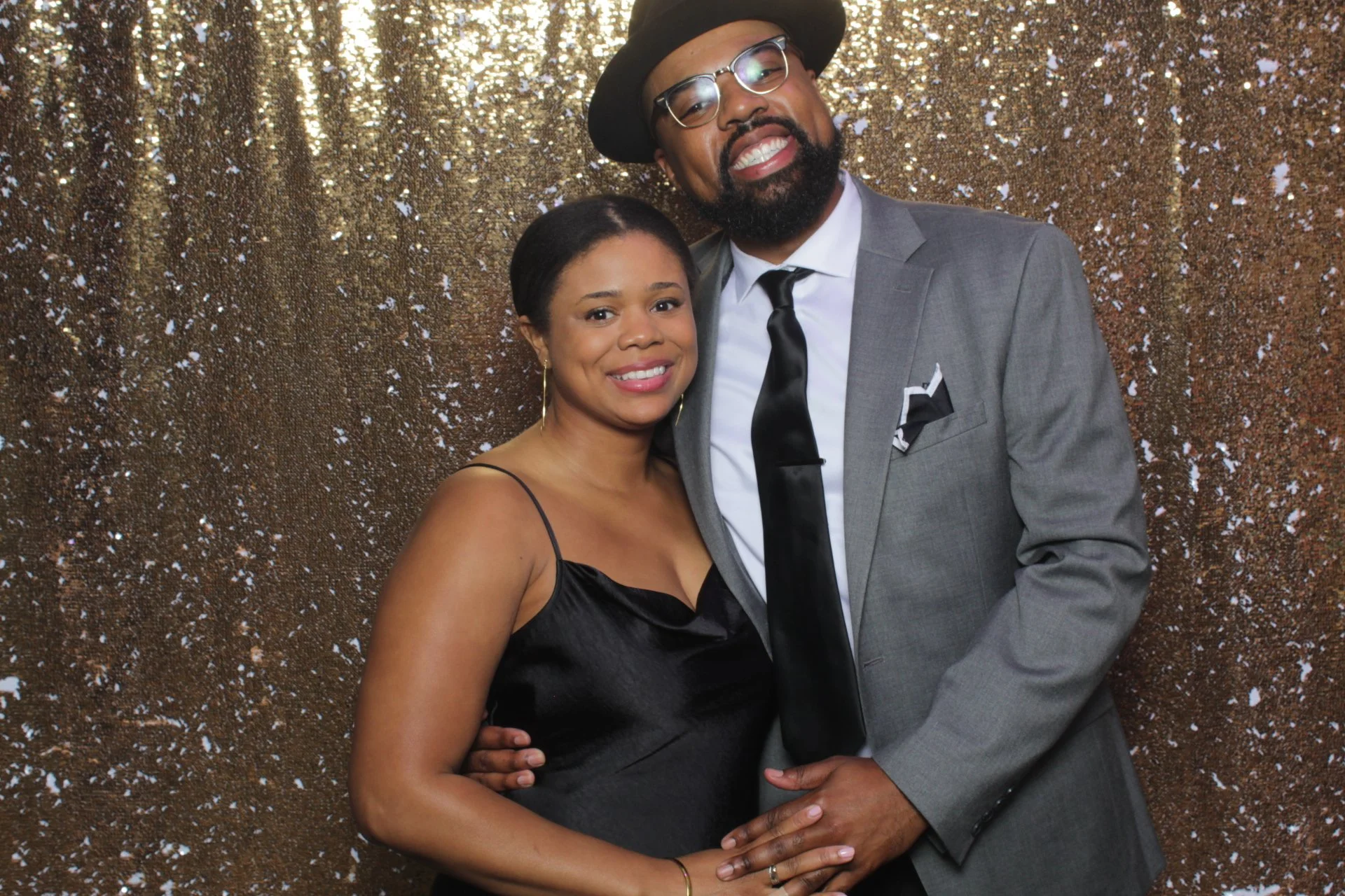A smiling woman in a black dress and a man in a gray suit, black tie, and black hat, standing close together against a glittery gold backdrop.