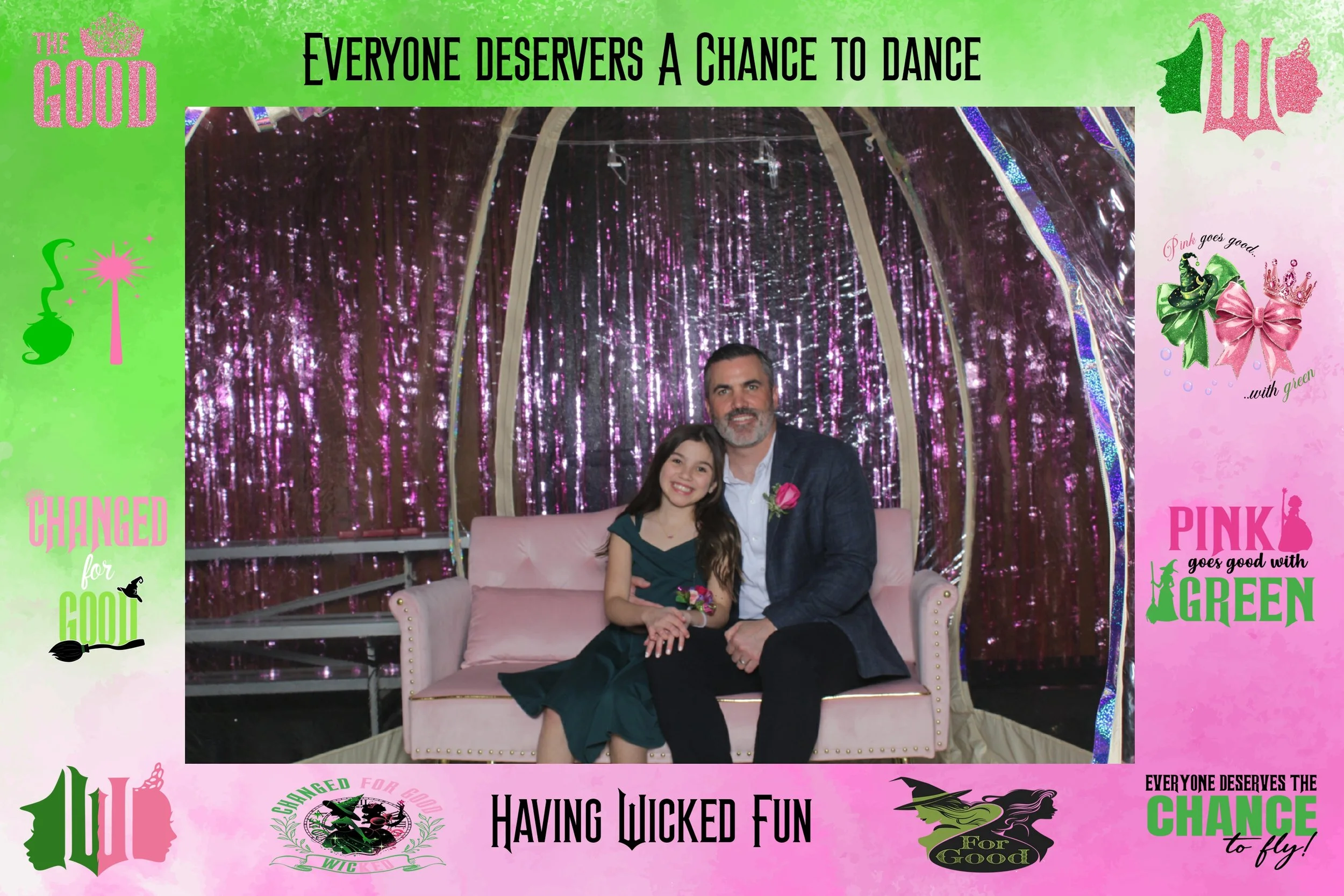 A man and a young girl sitting on a pink velvet sofa in front of a pink and purple shiny backdrop, surrounded by colorful banners and decorations with inspirational messages.
