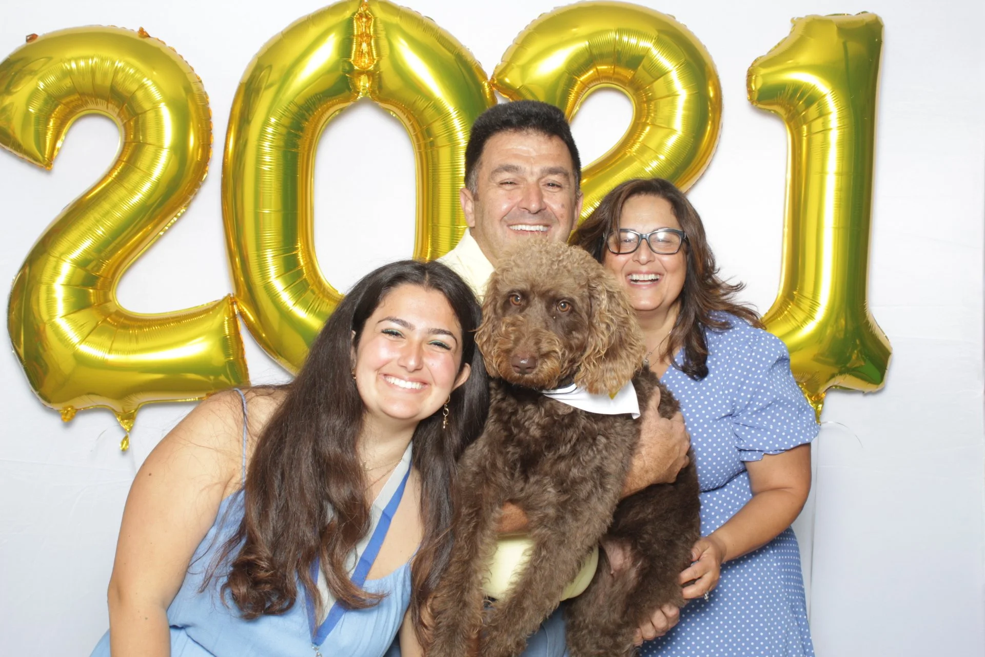 A group of four people, two women and two men, standing together and smiling, holding a brown curly-haired dog. Behind them are large gold balloons that spell out '2021' on a white wall, indicating a celebration or New Year's event.