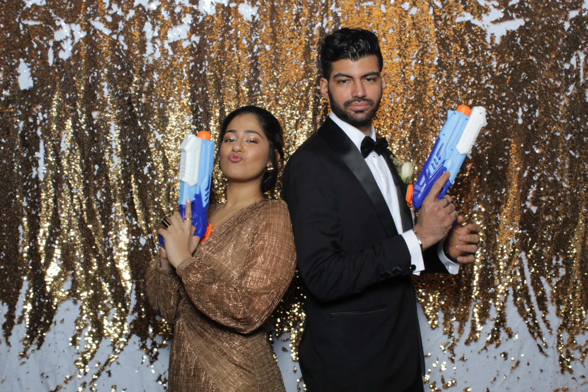 A couple dressed in formal attire posing back-to-back with toy water guns in front of a gold sequin backdrop.
