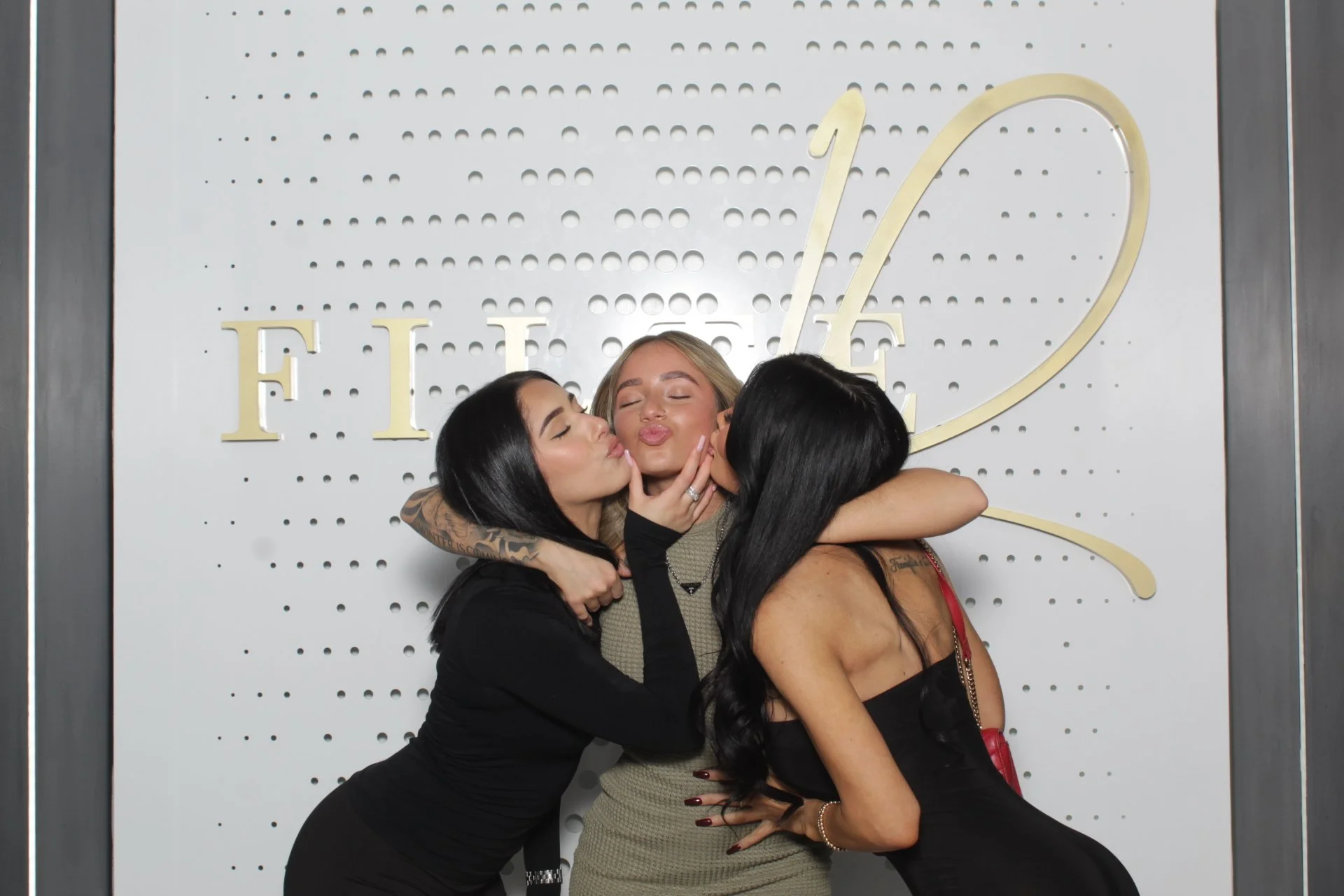 Three women sharing a hug and kisses in front of a decorative wall with the word 'LOVE' on it.