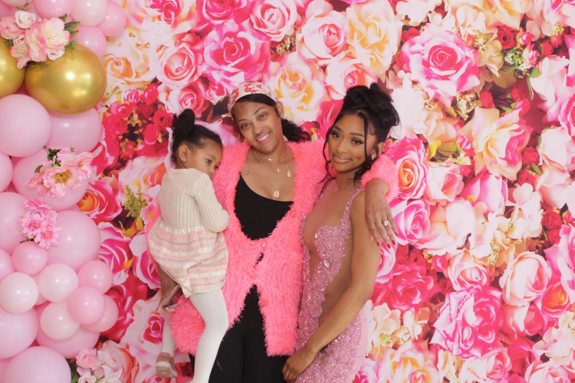 A family of three with two women and a young girl, standing in front of a pink floral backdrop with balloons.