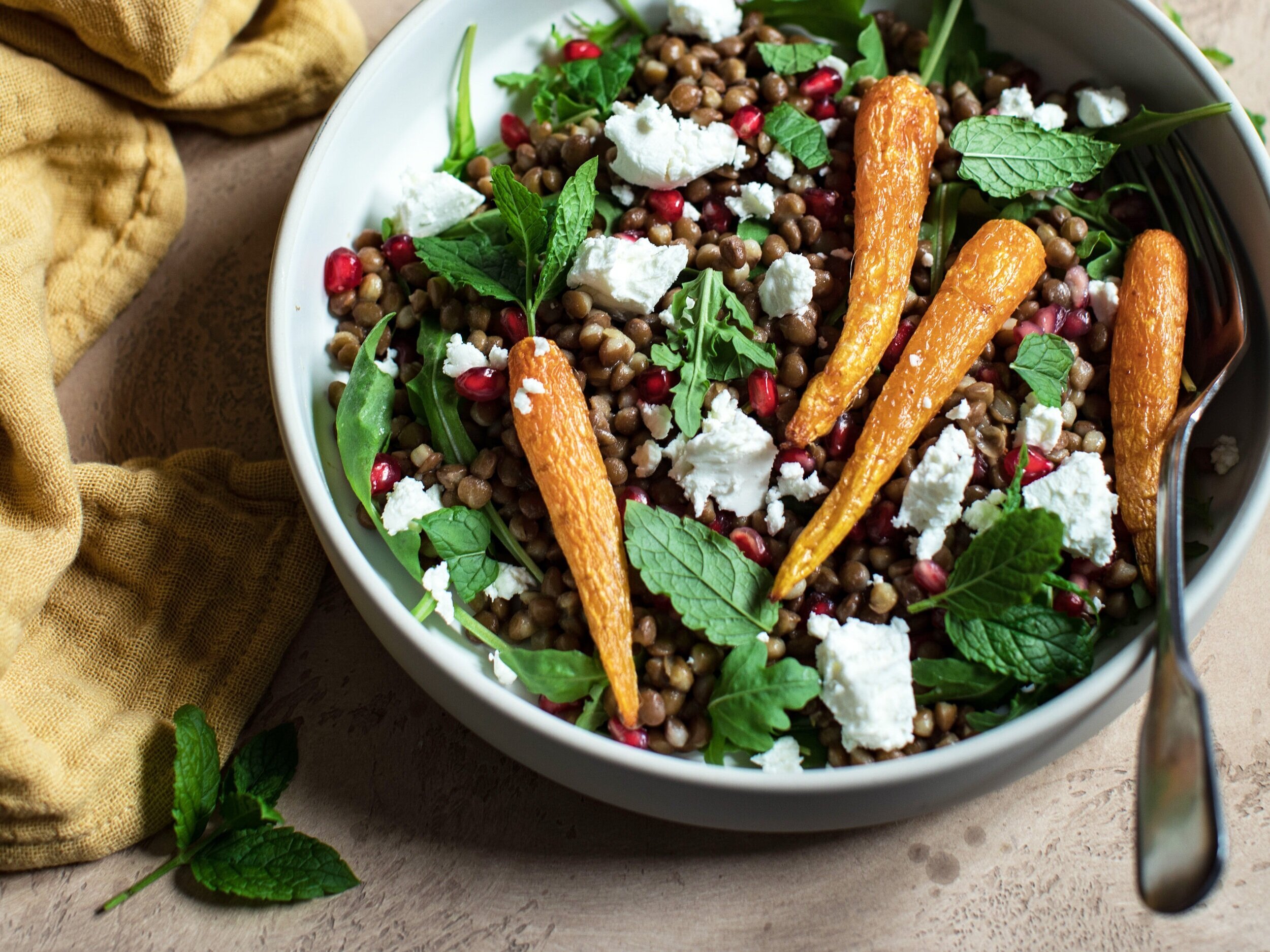  Round bowl with lentil salad. Crumbled feta, lentils, pomegranate seeds, roasted baby carrots and greens. Mustard dressing and tin of lentil top of image. 