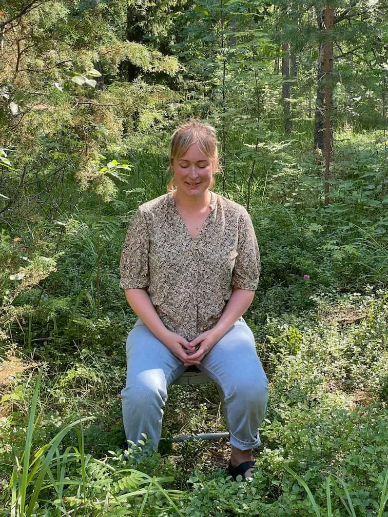 Elisa, Mindfulness teacher sitting down in a lush green forest smiling peacefully, eyes closed