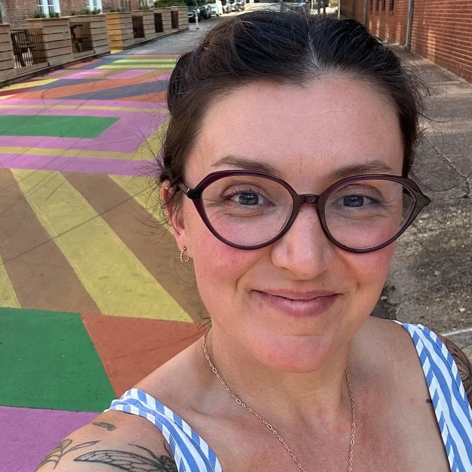 A woman wearing glasses and a blue and white striped top taking a selfie outdoors, standing on a colorful sidewalk with various painted patterns in pink, yellow, green, and brown, in a neighborhood with brick buildings and some trees in the background.