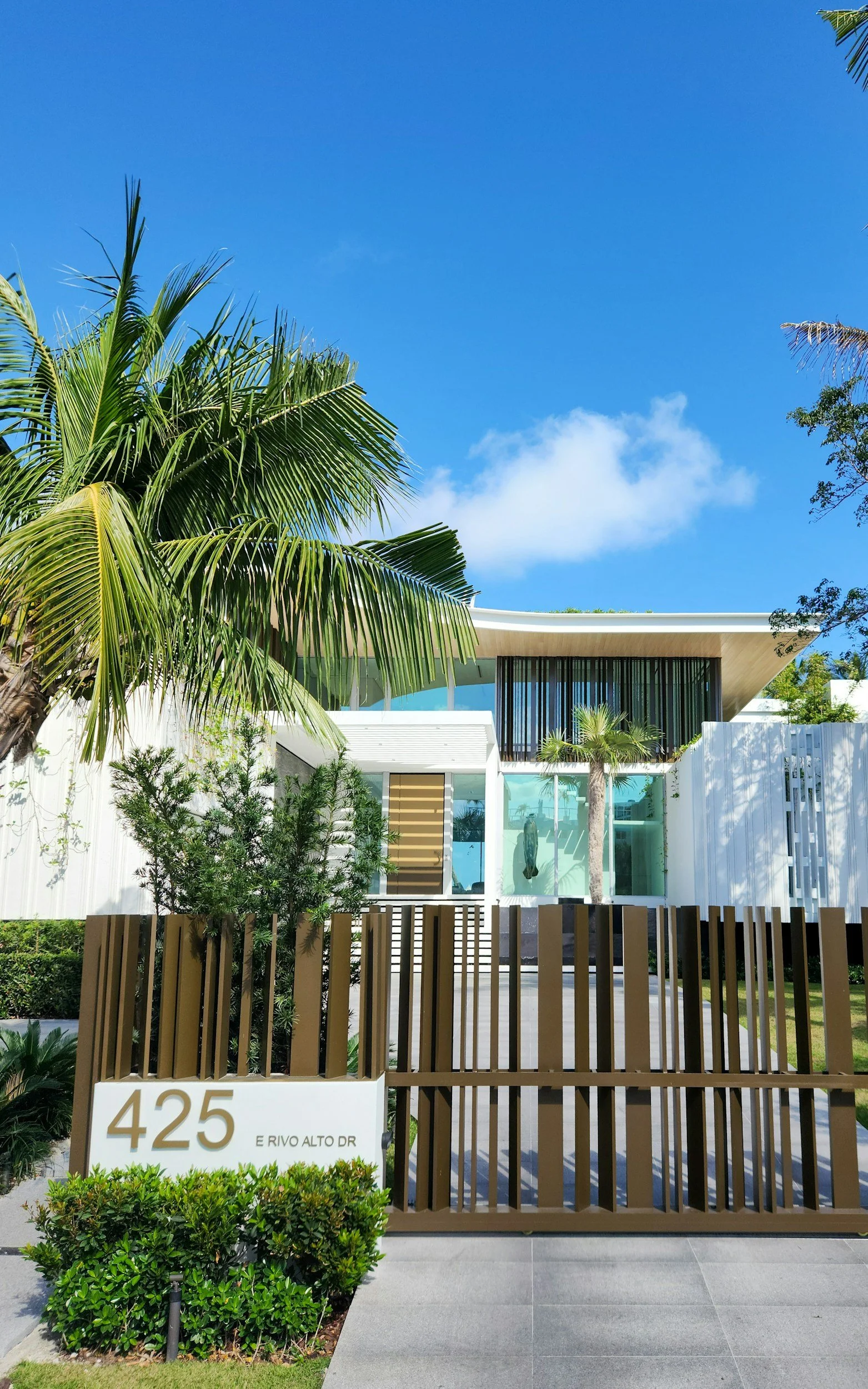 Modern white house with large glass windows surrounded by palm trees and greenery under a bright blue sky.