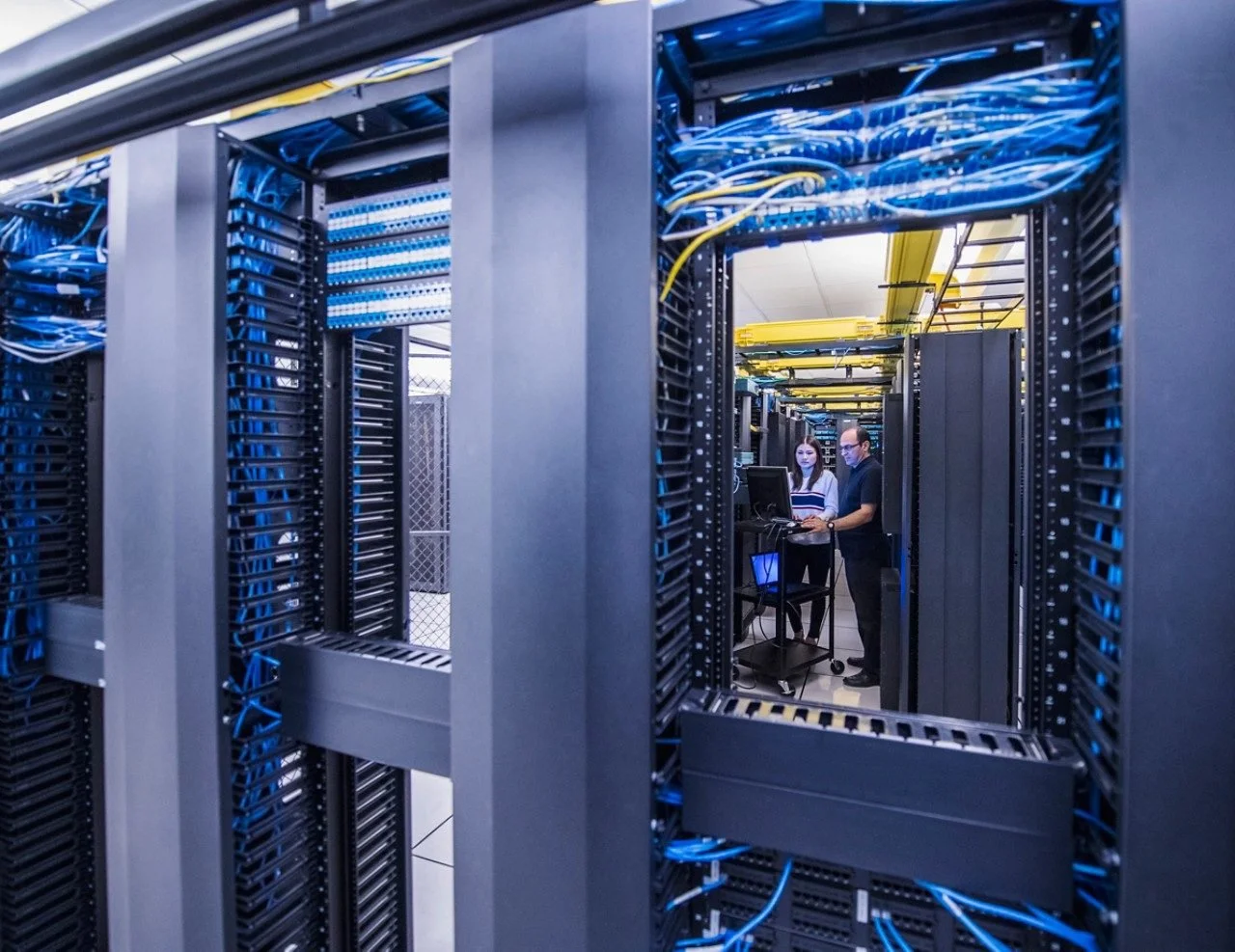 Two people in a data center working near server racks with blue and yellow cables, viewed through a server rack frame.