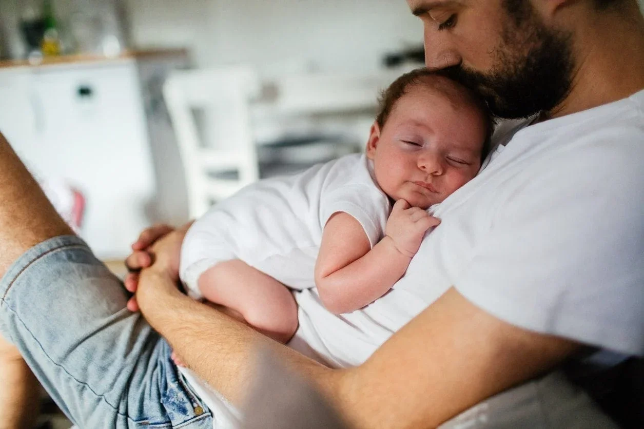 A man holding a sleeping baby close to his chest in a home setting.