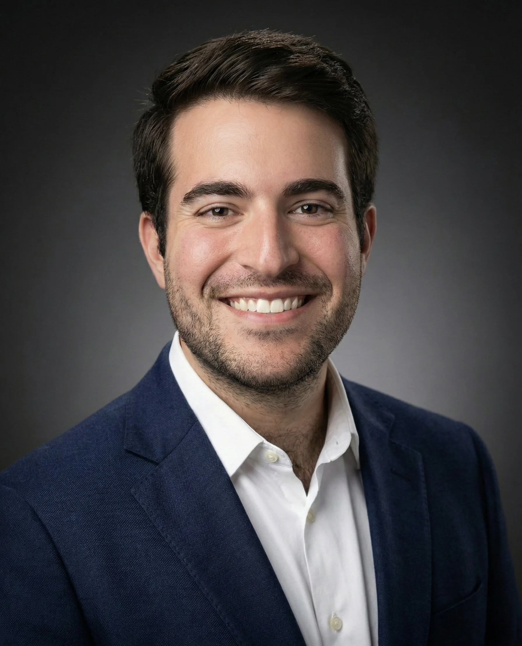 Professional headshot of a smiling man in a dark blue suit and white shirt, with dark hair and a short beard, against a dark gray background.