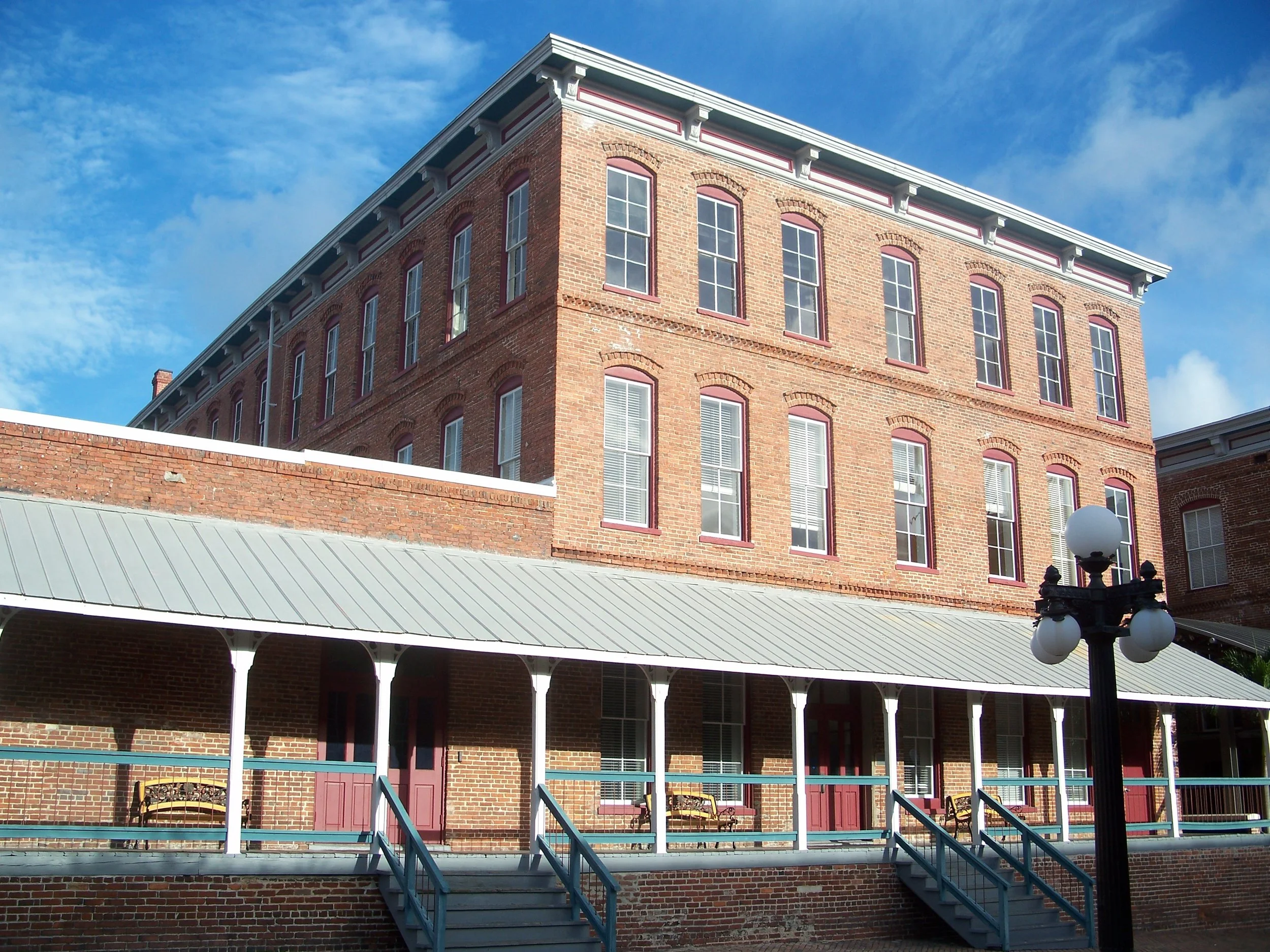 A historic red brick building with multiple stories and tall windows, with a metal-roofed porch and a black vintage streetlamp in front.