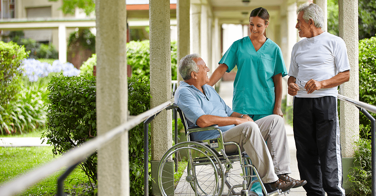 A caregiver talking to an elderly man in a wheelchair outside a building, with another elderly man standing nearby, surrounded by greenery.