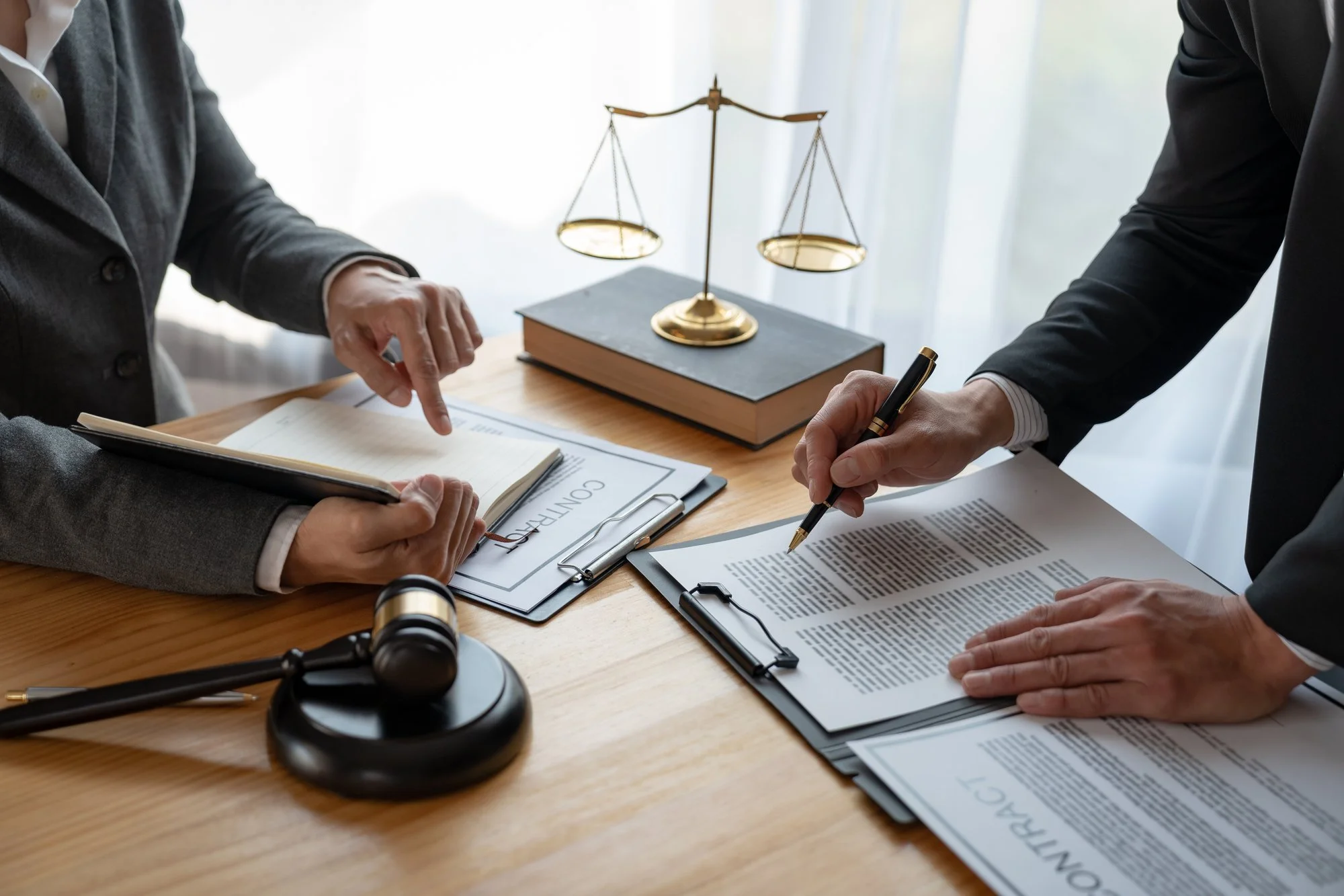 Two people discussing contracts at a desk, with legal documents, a gavel, and a balance scale visible.