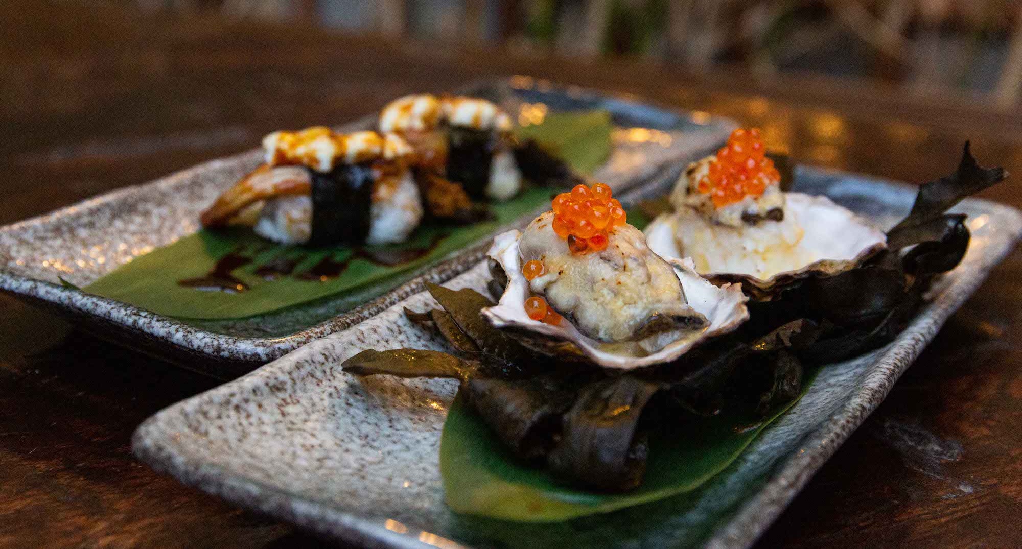 Grilled eel nigiri with torched goat’s cheese (back), and Grilled Oysters with Masago over Irish Seaweed and Banana Leaf