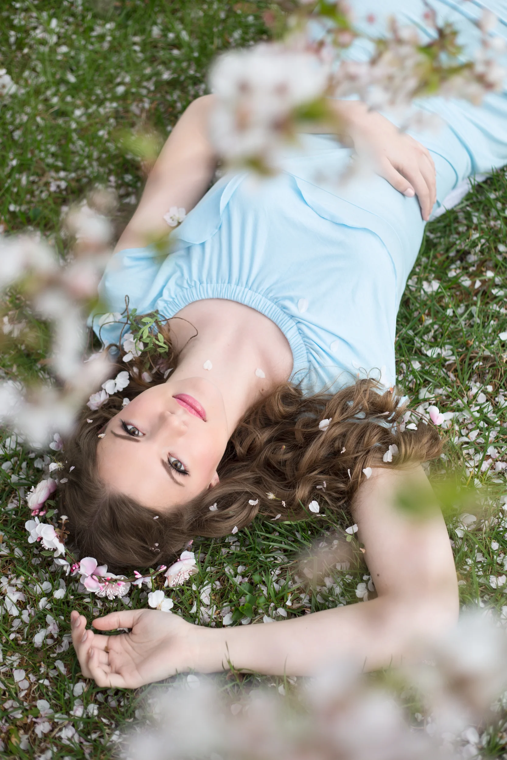 A young woman with long, curly brown hair lies on a grassy field surrounded by pink and white flowers, wearing a light blue dress with her eyes open.