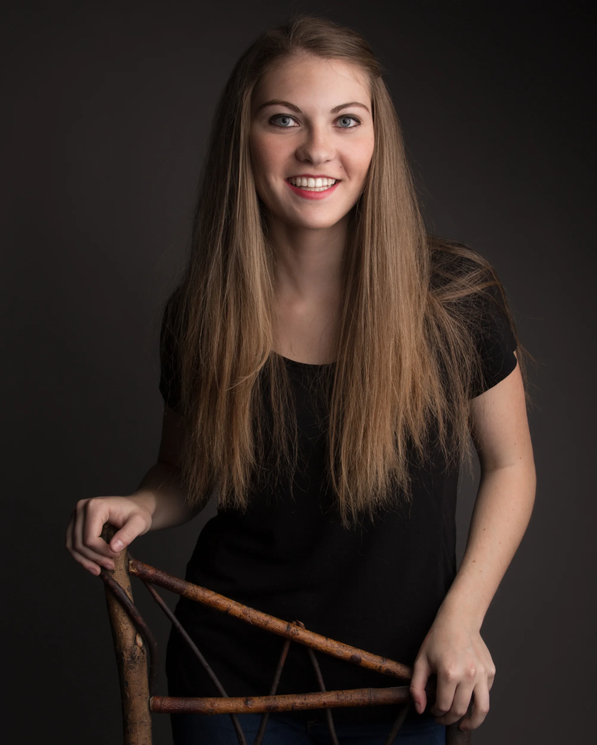 Young woman with long straight hair smiling, holding a wooden chair, wearing a black shirt, dark background.