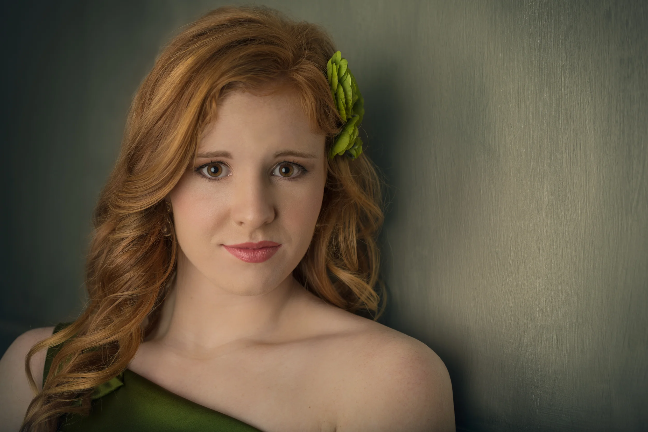 A young woman with red hair decorated with green flowers, looking at the camera against a dark green background.