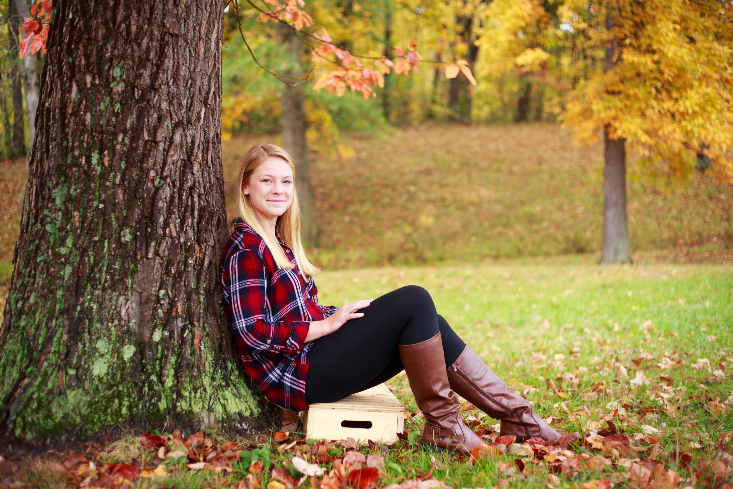 A young woman with blonde hair sitting against a large tree in a fall park, wearing a red plaid shirt, black leggings, and brown boots, with autumn leaves on the ground and colorful trees in the background.