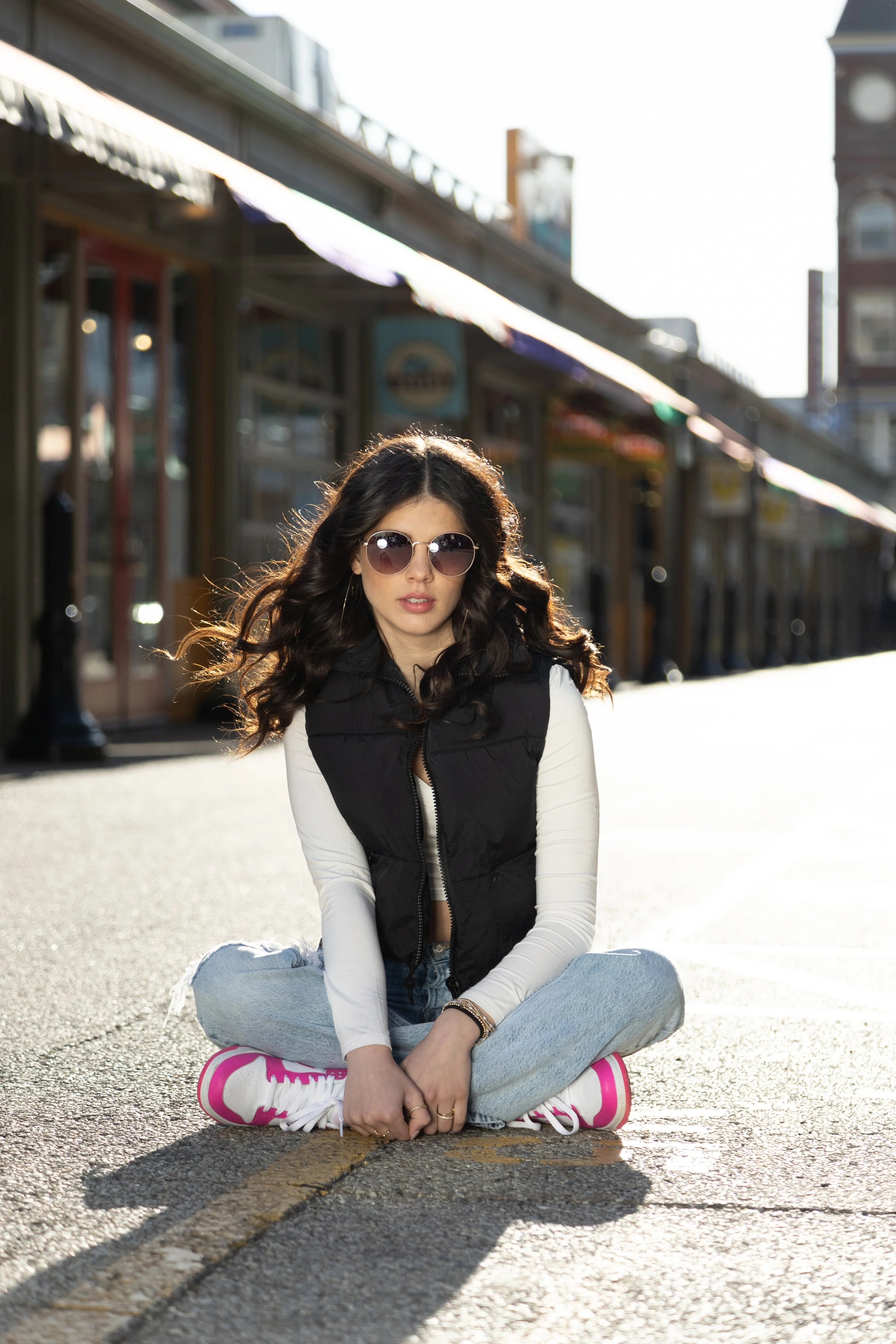A woman sitting cross-legged on the street in front of storefronts, wearing sunglasses, a black vest, white long-sleeve shirt, light jeans, and pink sneakers.