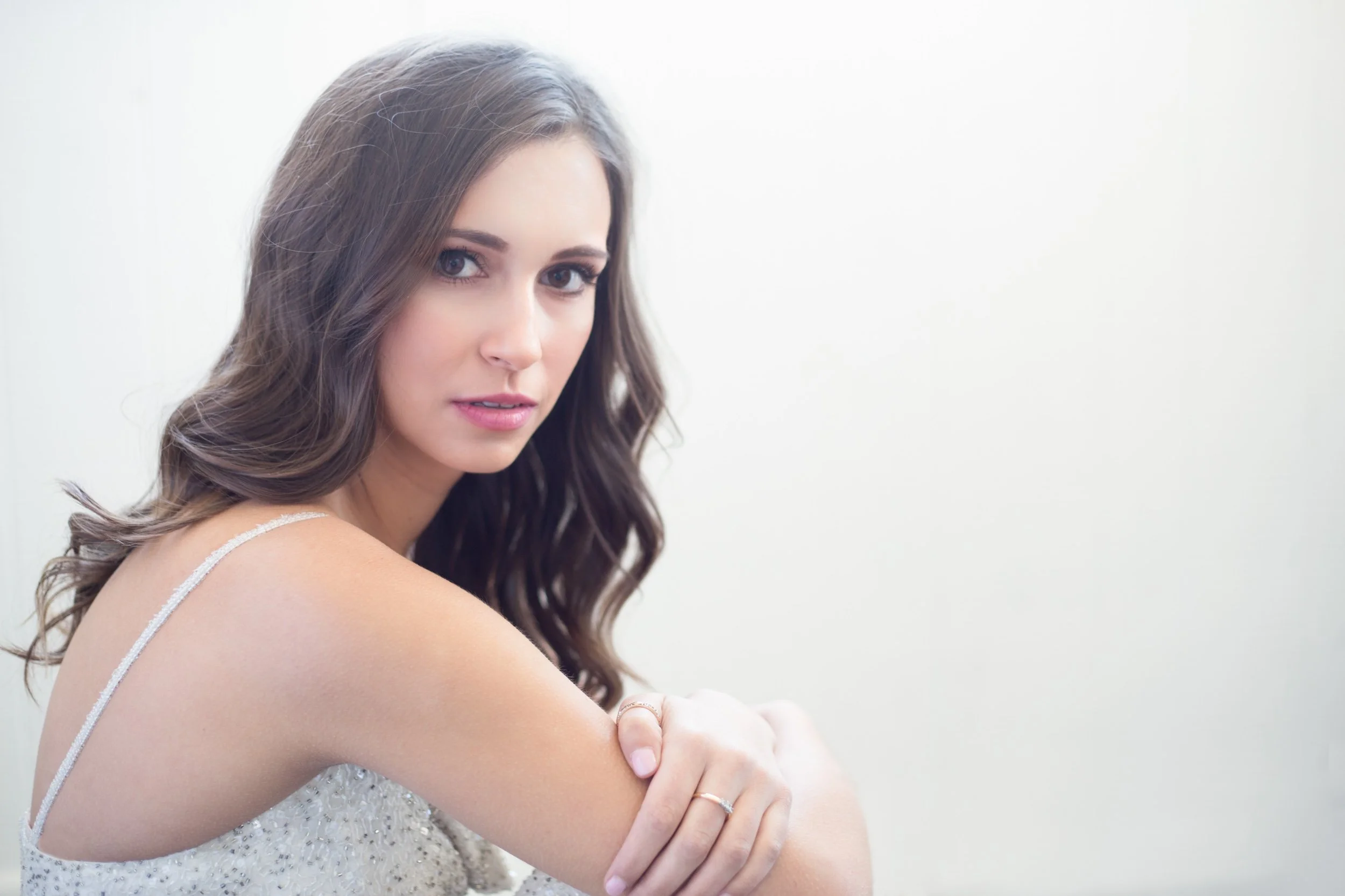 A young woman with long, wavy brown hair and blue eyes looking at the camera, wearing a sleeveless top, with her arm crossed and a ring on her finger, against a plain white background.