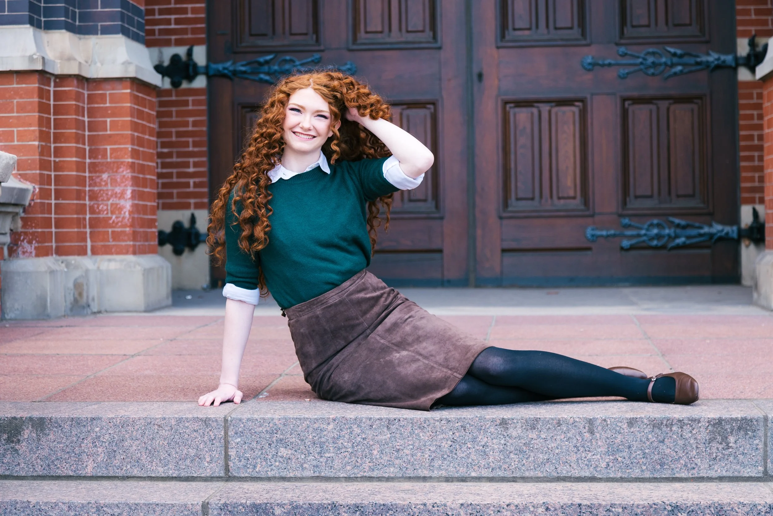 A young woman with long curly red hair sitting on stairs in front of a large wooden door, smiling and touching her hair.