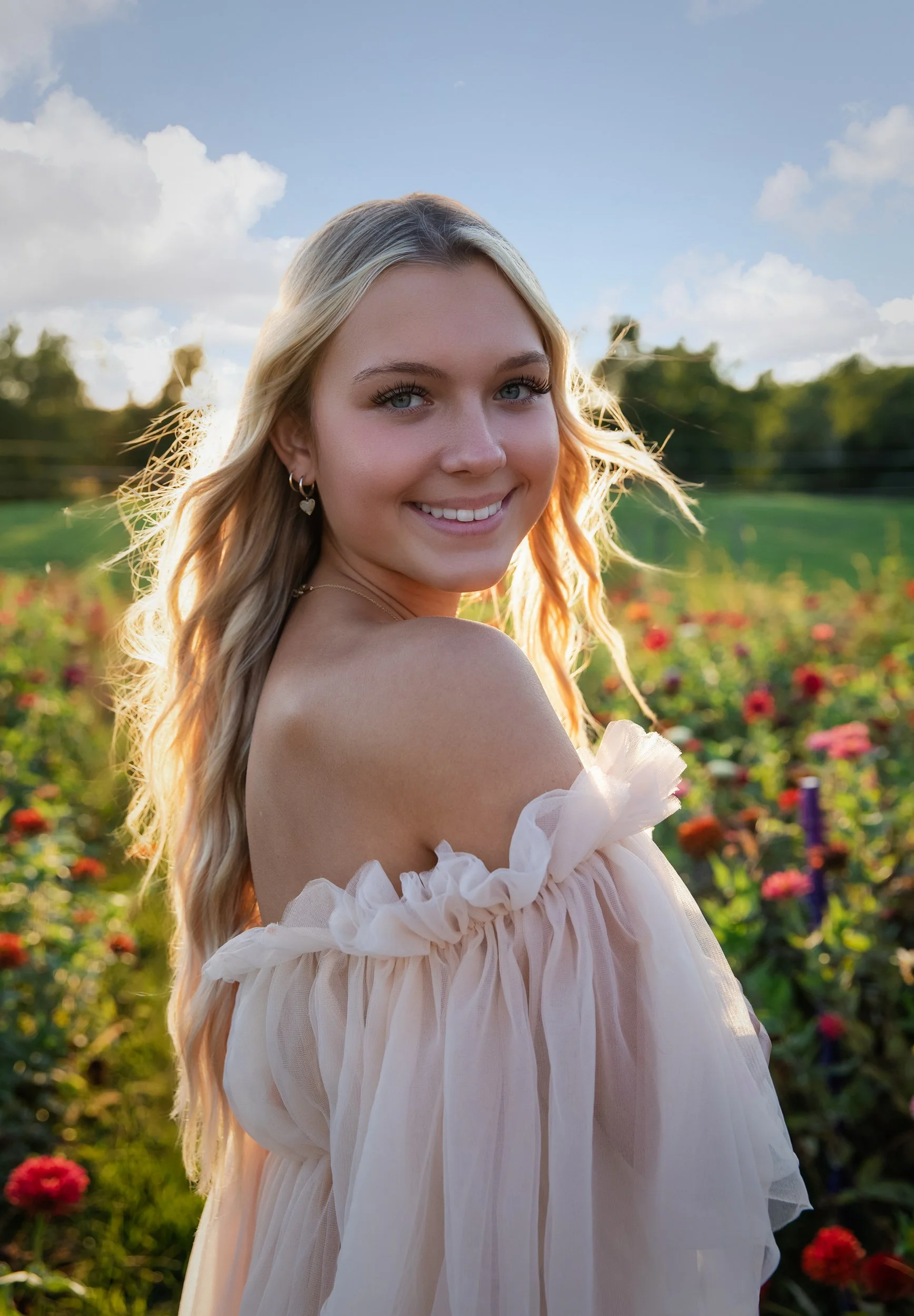 A young woman with blonde hair and blue eyes smiling outdoors in a field of colorful flowers during sunset.
