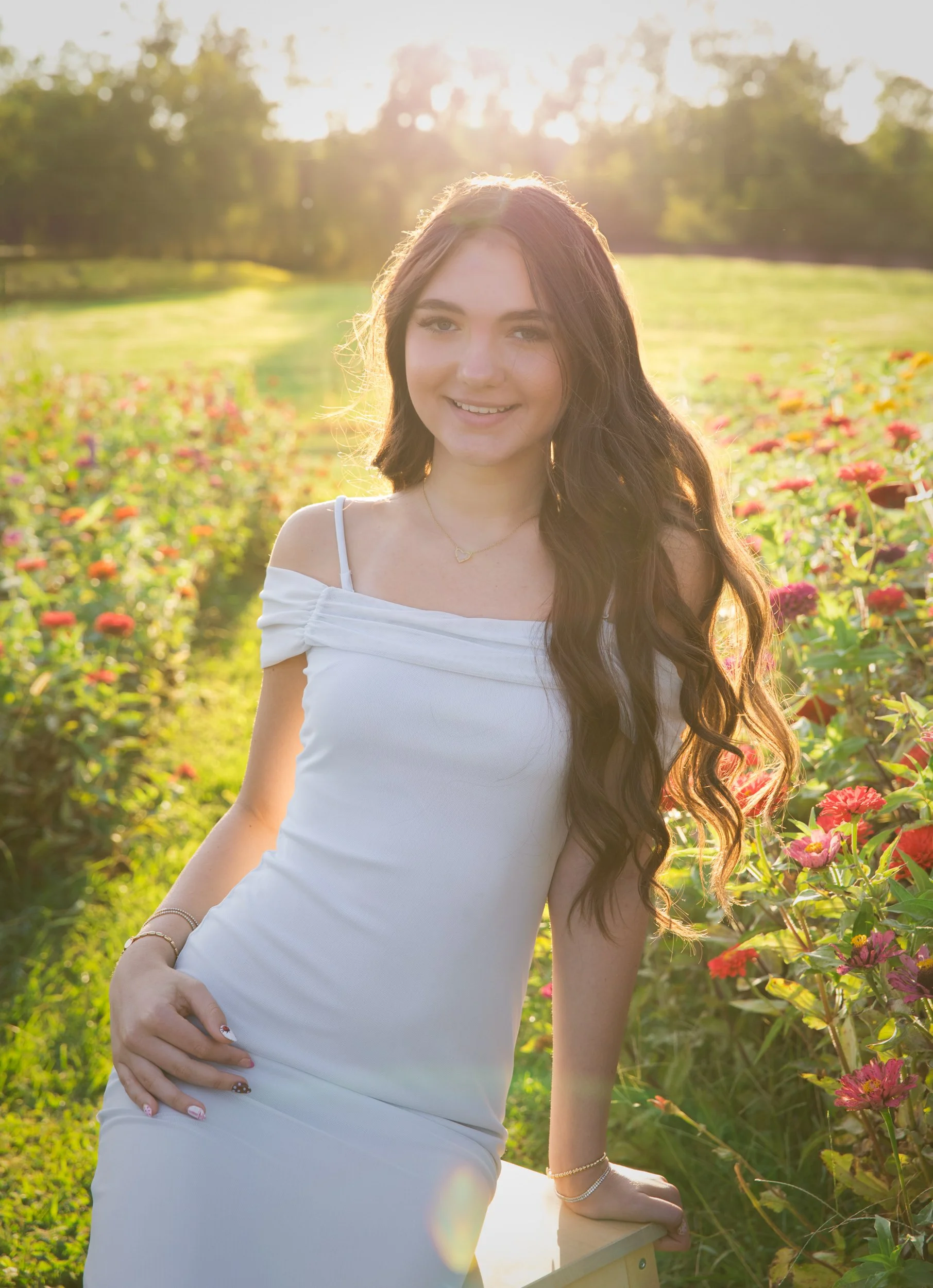 A young woman in a white dress sitting on a yellow bench in a flower field during sunrise or sunset, with long wavy hair and a smile.
