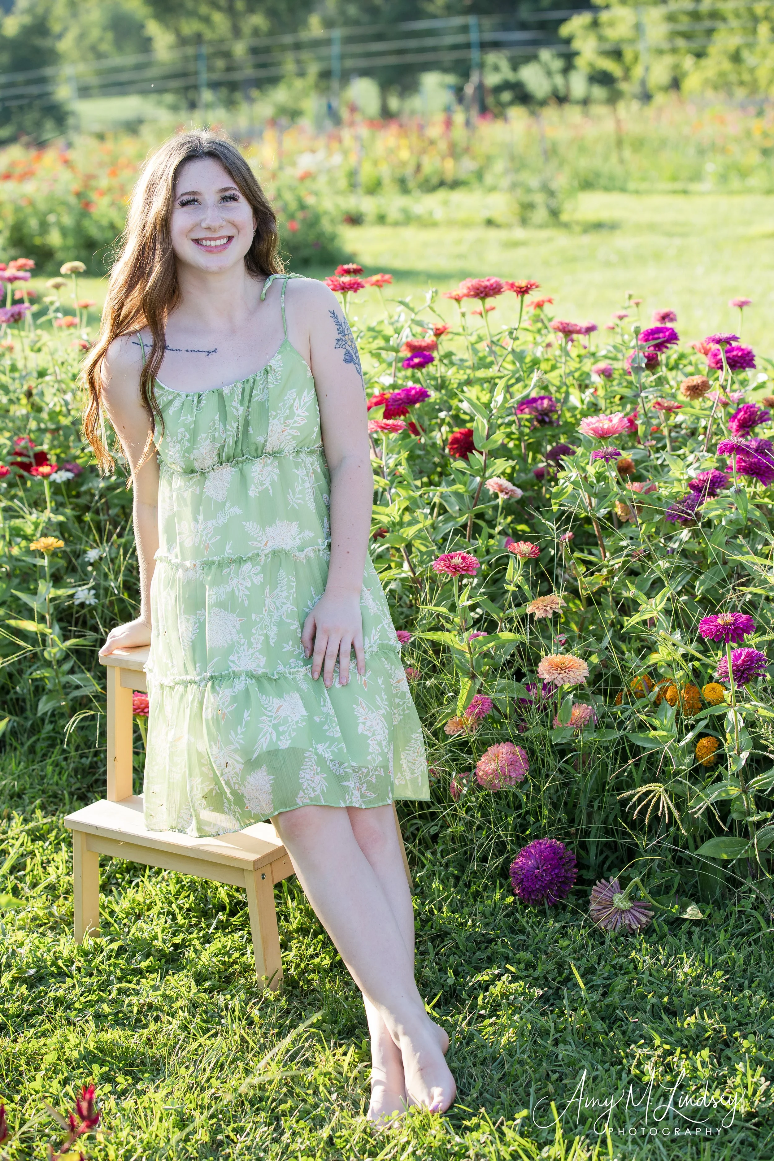 Woman in a green floral dress sitting on a small wooden stool in a colorful flower garden, smiling at the camera.