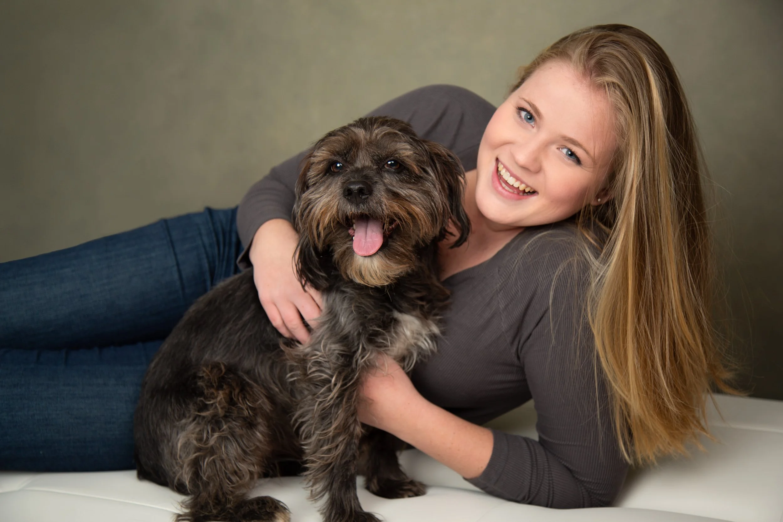 A woman with long blonde hair, smiling, lying on her side on a light-colored surface, hugging a black and brown fluffy dog with an open mouth and tongue out. The background is a plain, muted wall.