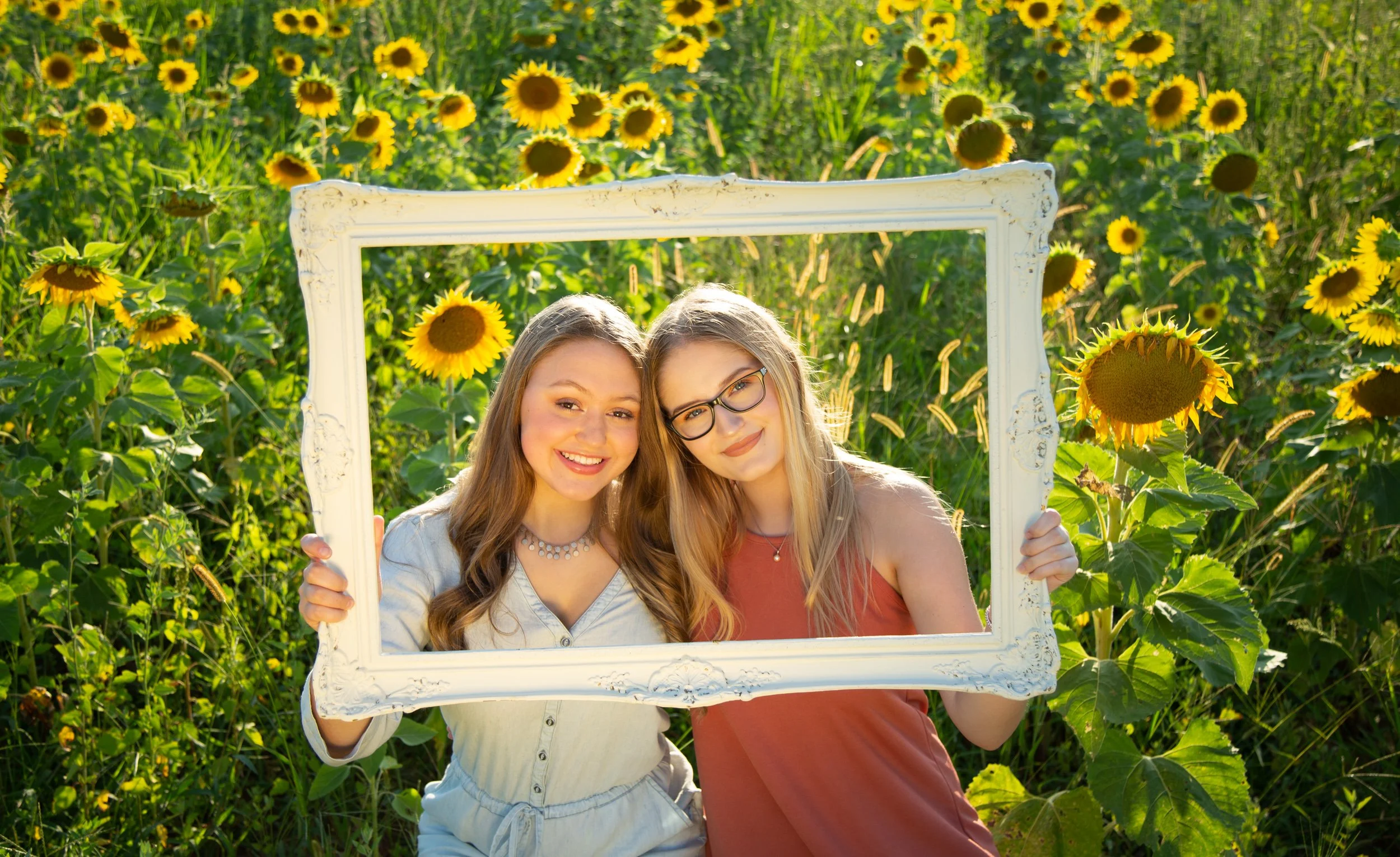 Two smiling young women holding a white picture frame in a sunflower field during daytime.