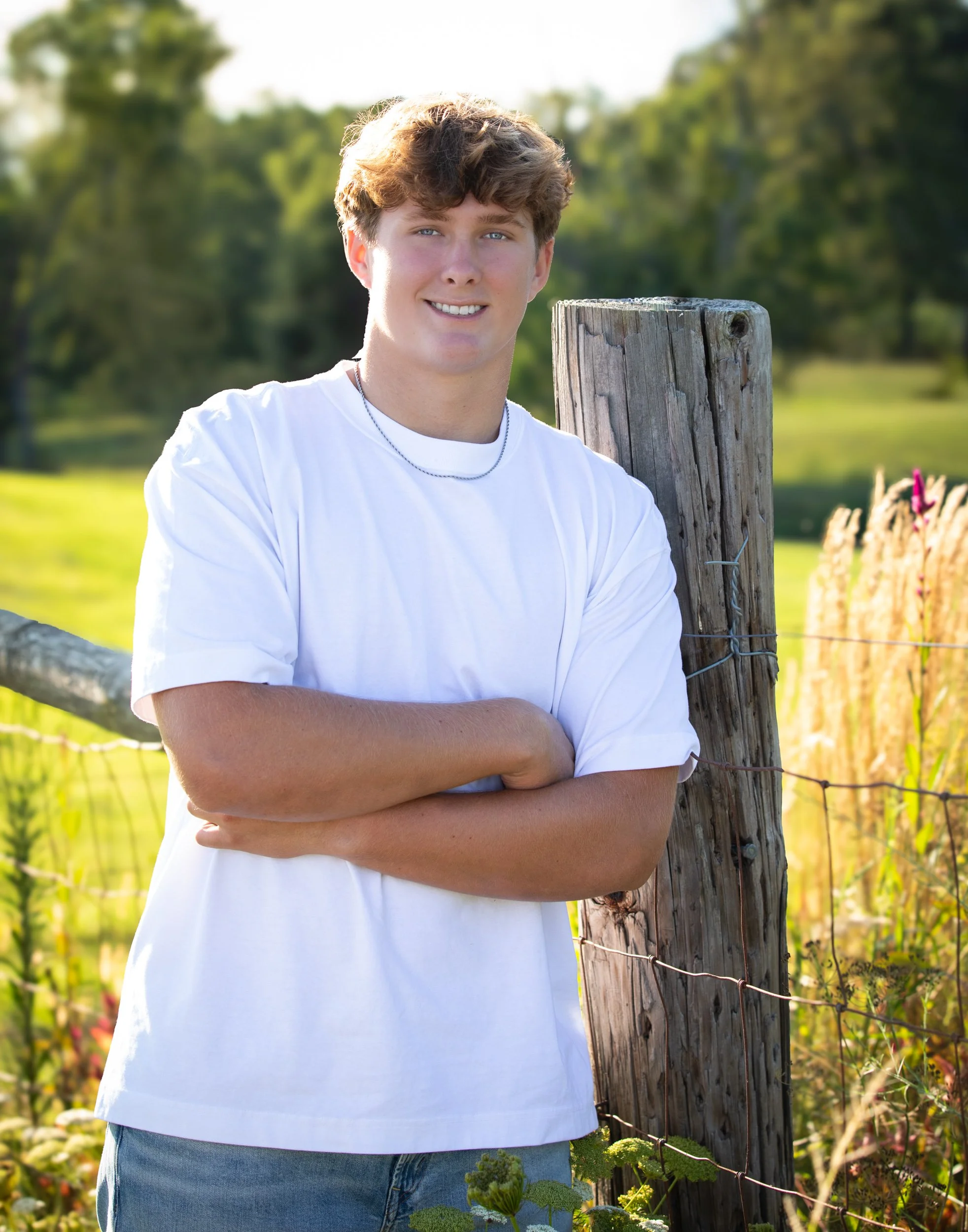 A young man with curly hair and blue eyes stands outdoors on a sunny day, smiling with arms crossed, near a wooden post and barbed wire fence in a field with green trees and grass in the background.