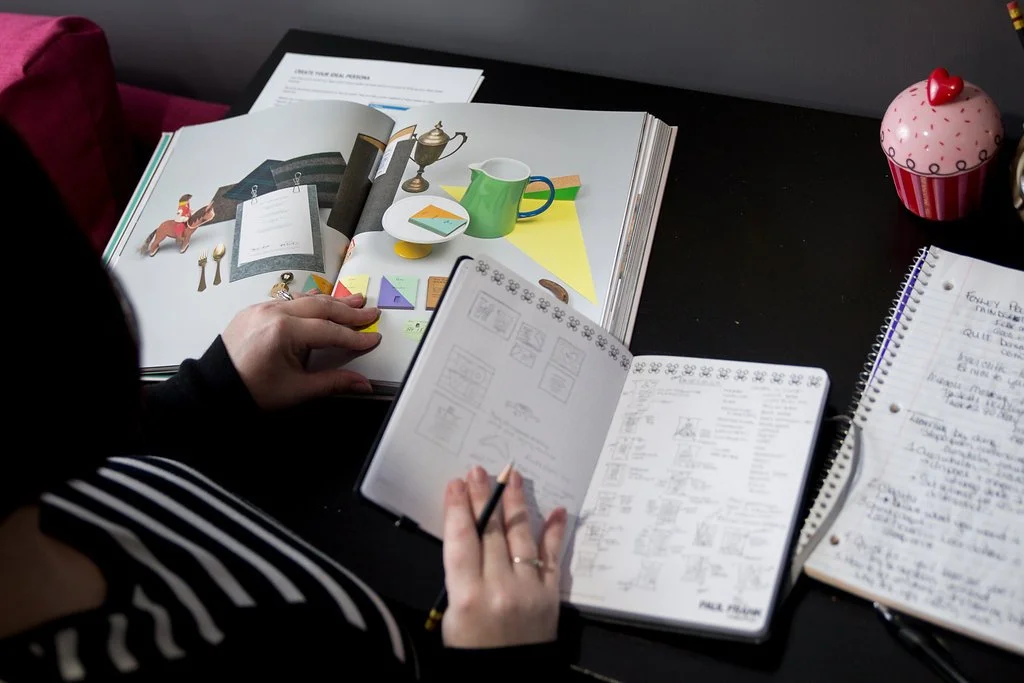 Angie looking through design book with sketchbook open on desk. A notebook is close by with notes.